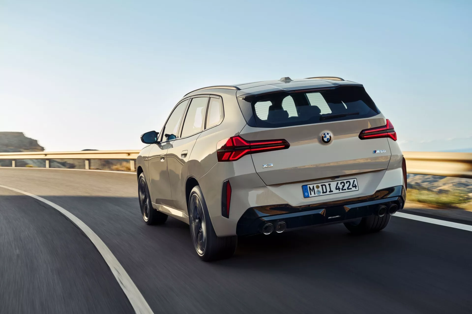 Obraz przedstawia BMW X3 2024 (G45) z tylnego trzech czwartych widoku, podkreślając zarówno boczne, jak i tylne profile, gdy wydaje się być w ruchu na drodze z widocznymi oznaczeniami pasów. Samochód ma charakterystyczne LED-owe światła tylne, czujniki wspomagające parkowanie na tylnym zderzaku, podwójny układ wydechowy, tylny dyfuzor i wieloszprychowe felgi aluminiowe. Widoczne są również lusterka boczne, prawdopodobnie wyposażone w zintegrowane kierunkowskazy i zaawansowane funkcje. Design i elementy technologiczne pojazdu odzwierciedlają nowoczesne osiągnięcia w motoryzacji.