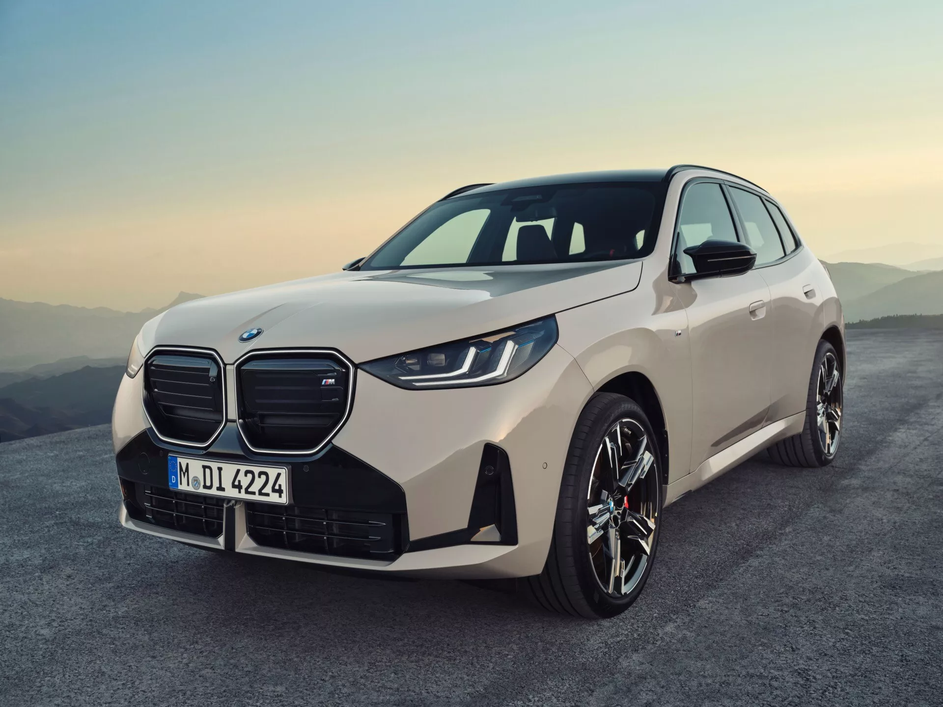 Perspektywa z trzech czwartych przodu BMW X3 2024 (G45) ukazująca charakterystyczny podwójny grill, eleganckie reflektory LED, zintegrowane światła przeciwmgielne, sportowe felgi aluminiowe i wyrafinowany jasnobeżowy kolor pod zmierzchowym niebem z ciepłymi odcieniami. Pojazd jest lekko wychylony w lewo, podkreślając swoje aerodynamiczne kontury i nowoczesny design.