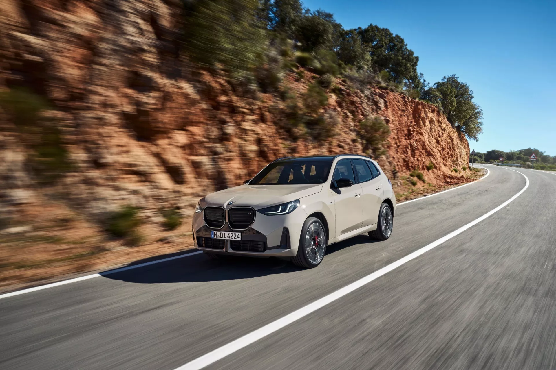 Dynamiczne ujęcie BMW X3 2024 (G45) poruszającego się po krętej drodze w świetle dziennym. Uchwycony pod kątem trzy czwarte przodu, obraz ukazuje aerodynamiczny, elegancki kształt samochodu, wyraźny przód grilla, reflektory LED/laserowe i wieloszprychowe koła. Lusterka boczne mają wbudowane kierunkowskazy. Jasny, neutralny kolor kontrastuje z naturalnym, skalistym tłem, co sugeruje przystosowanie pojazdu do różnych terenów. Rozmycie ruchu na kołach i tle podkreśla prędkość.