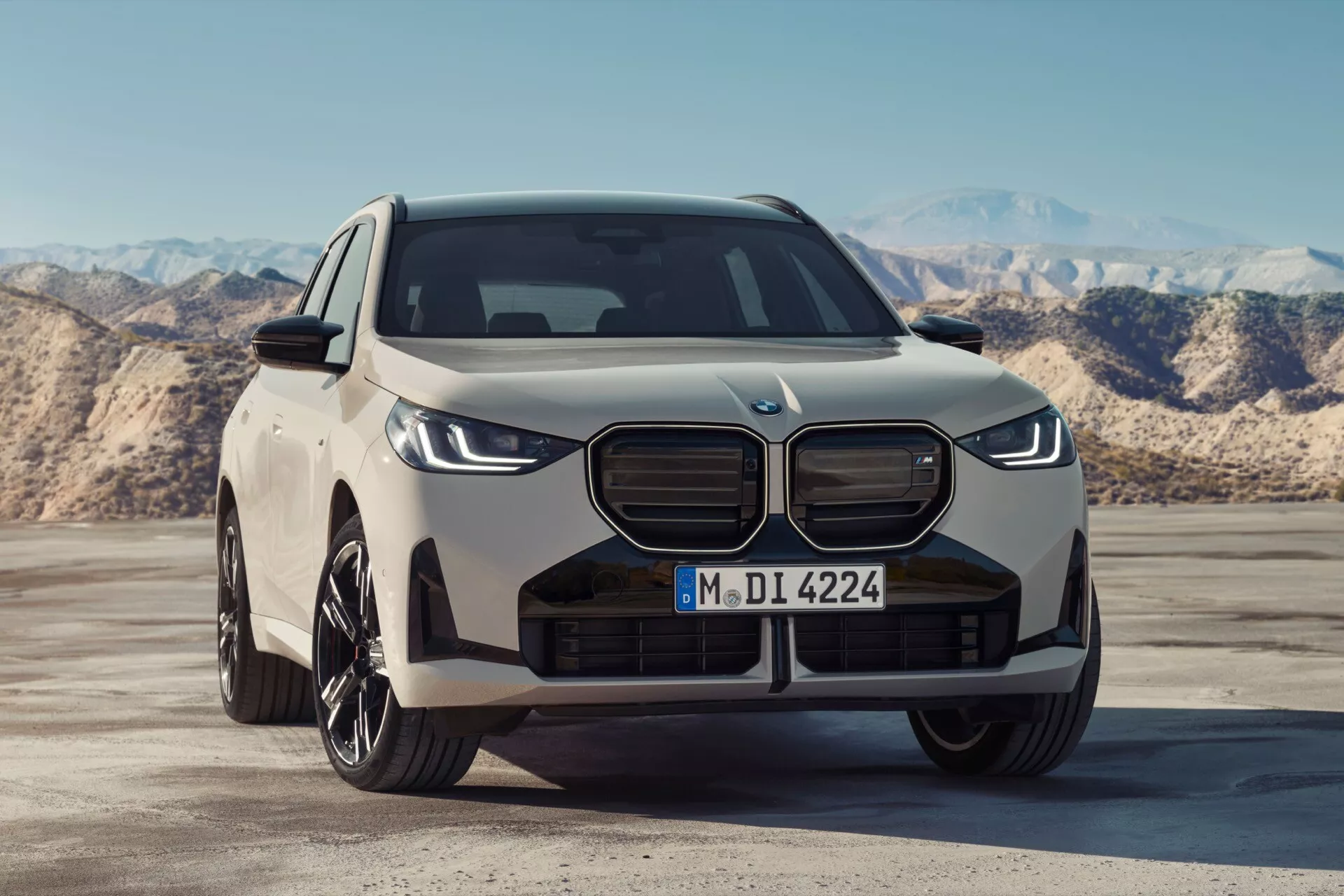 To zdjęcie przedstawia przedni trójkątny widok SUV-a BMW X3 2024 (G45) z lekko niskiej perspektywy, ukazując kratkę przednią, zderzak, maskę i część bocznego profilu. Pojazd cechuje się charakterystycznymi dla marki dużymi, pionowo wydłużonymi kratkami nerkowymi, prawdopodobnie zasłoniętymi, co może sugerować elektryczny lub hybrydowy napęd, nowoczesnymi, smukłymi reflektorami oraz sportowymi, wieloramiennymi felgami z lekkiego stopu. Centralny wlot powietrza, ostre linie maski, duży emblemat oraz lusterka z wbudowanymi kierunkowskazami podkreślają futurystyczny i agresywny wygląd pojazdu.