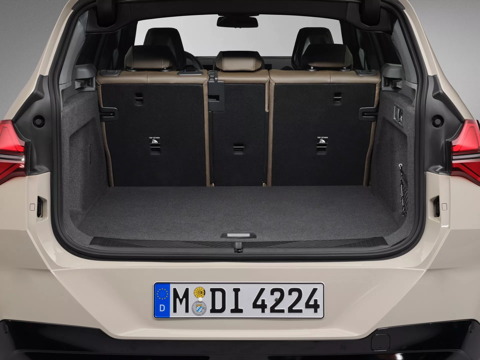 Wnętrze tylne BMW X3 2024 (G45) widziane bezpośrednio z tyłu z otwartą klapą bagażnika, ukazujące przestronną przestrzeń ładunkową z płaską podłogą i częściowo złożonymi tylnymi siedzeniami w konfiguracji 60/40. Bagażnik posiada dywanową podłogę, oparcia siedzeń z tkaniny lub skóry oraz plastikowe wykończenia. Oświetlenie jest profesjonalne, podkreślające szczegóły bez ostrych cieni. Widoczna jest europejska tablica rejestracyjna, co sugeruje europejski model. Na zdjęciu nie widać żadnych rozpoznawalnych emblematów marki ani modelu.