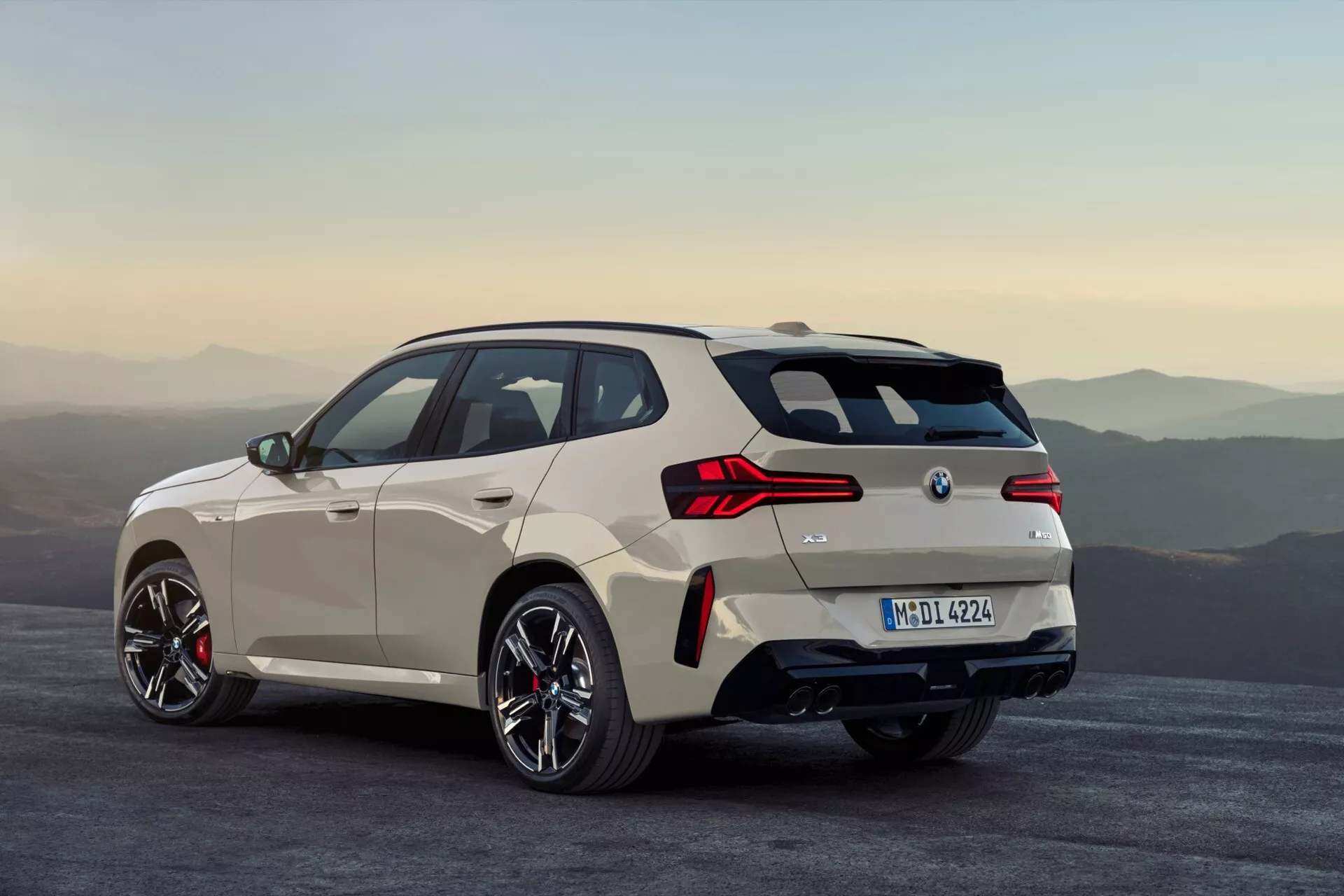 Obraz nowoczesnego hatchbacka, BMW X3 2024 (G45), ujęty pod kątem trzech czwartych od tyłu. Samochód, lekko przesunięty w prawo, wydaje się zaparkowany na szerokiej, płaskiej powierzchni z rozmazanym górskim tłem. Jasne nadwozie kontrastuje dobrze z otoczeniem, ukazując felgi wieloszprychowe, niskoprofilowe opony i duże zaciski hamulcowe. Eleganckie linie aerodynamiczne, pochylony dach, charakterystycznie zaprojektowane tylne światła, widoczne tylne czujniki, dyfuzor, podwójny układ wydechowy, subtelny tylny spojler, antena w kształcie płetwy rekina na dachu i składane boczne lusterka podkreślają nacisk samochodu na osiągi i zaawansowane technologie.