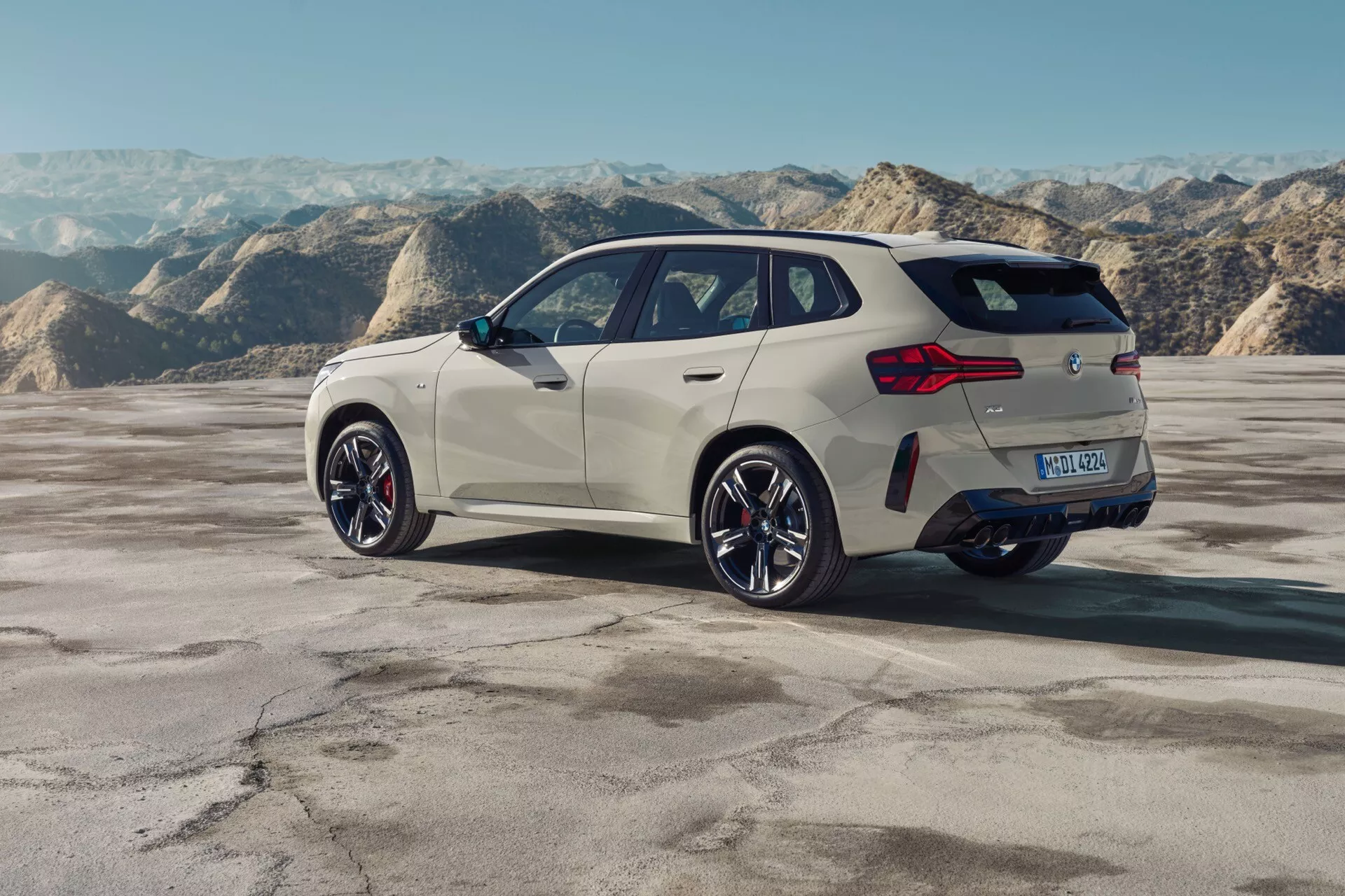 Widok z tyłu pod kątem trzy czwarte na BMW X3 2024 (G45) pokazujący prawą stronę pojazdu. Obrazek podkreśla tylną część, część bocznego profilu, design tylnych świateł, boczne okna i tylne koło. Widoczne są charakterystyczne elementy, takie jak rzeźbiony tylny zderzak, tylny spojler, podwójny układ wydechowy i stylowe wieloramienne felgi aluminiowe. Samochód, pomalowany na jasny kolor, taki jak biały lub kremowy, kontrastuje z ciemniejszymi elementami tylnymi dyfuzorami, listwami okiennymi i felgami aluminiowymi. Pojazd znajduje się w plenerze, na płaskim terenie z górzystym tłem, co może sugerować pustynię lub suche jezioro. Technologia LED w klastrach świetlnych, zaawansowane materiały aluminiowe w kołach, aerodynamiczne cechy nadwozia i widoczne czujniki lub kamery sugerują zaawansowane systemy wspomagania kierowcy. Tablica rejestracyjna została rozmazana dla prywatności.