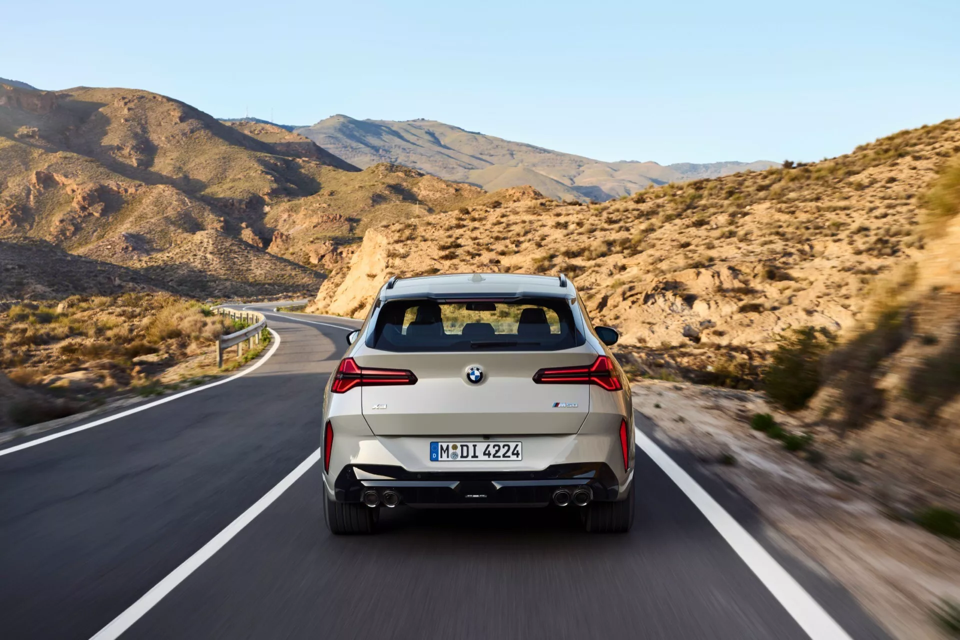 Widok tylni poruszającego się BMW X3 2024 (G45) na dwupasmowej drodze otoczonej górzystym terenem, z nowoczesnymi światłami tylnymi, podwójnym układem wydechowym i europejską tablicą rejestracyjną z niemiecką rejestracją.