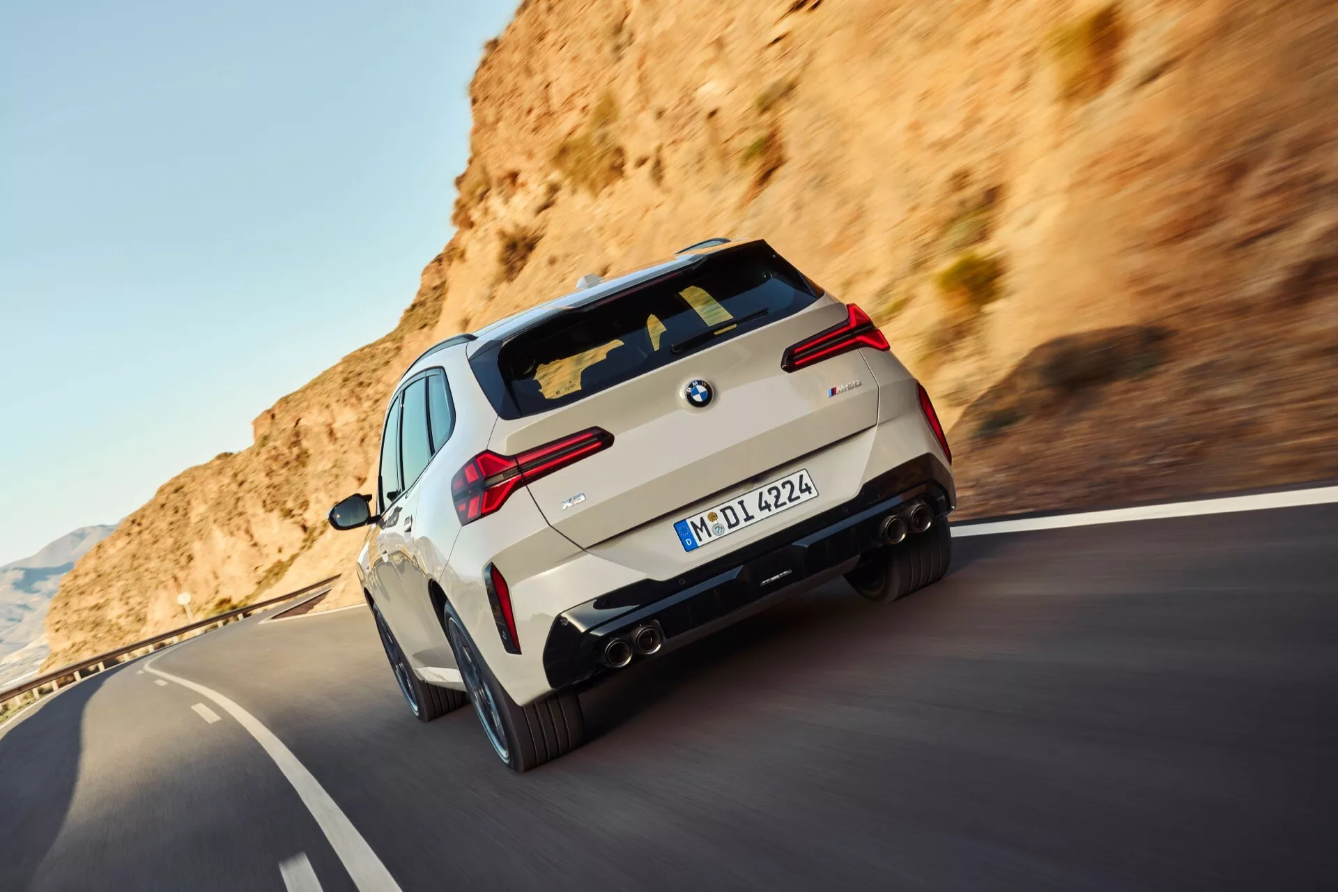 Dynamiczne ujęcie pod kątem tylnym poruszającego się BMW X3 (G45) 2024 na górskiej drodze. Samochód uchwycony w świetle dziennym, podkreślający typowe cechy designu BMW, takie jak widoczny badge, tylne światła, zderzak, tylne okno i sportowe felgi wieloramienne. Tablica rejestracyjna i oznaczenie 'M' sugerują wysokowydajny model z dywizji M BMW. Rozmycie ruchu na kołach i drodze oddaje poczucie prędkości i ruchu.