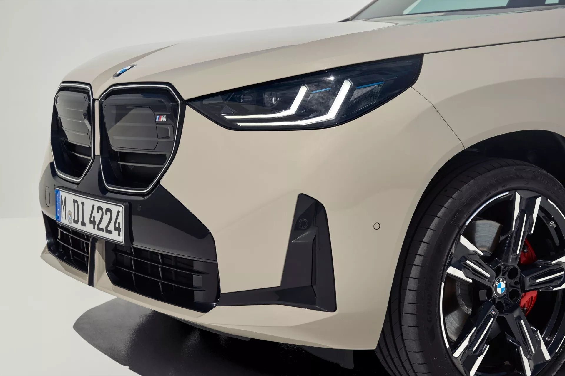 Zbliżenie na BMW X3 2024 (G45) z przedniego lewego kąta, prezentując wyraźną szeroką osłonę chłodnicy nerkowatego kształtu, eleganckie reflektory LED, częściowo widoczną maskę i przedni zderzak z wlotami powietrza. Sportowy design obejmuje wieloszprychowe alufelgi z czerwonymi zaciskami hamulcowymi i tablicę rejestracyjną 'M DL 4224', co sugeruje rejestrację w Monachium. Podkreślono zaawansowaną aerodynamikę i technologię pojazdu, przedstawiając dynamiczny i luksusowy wygląd.