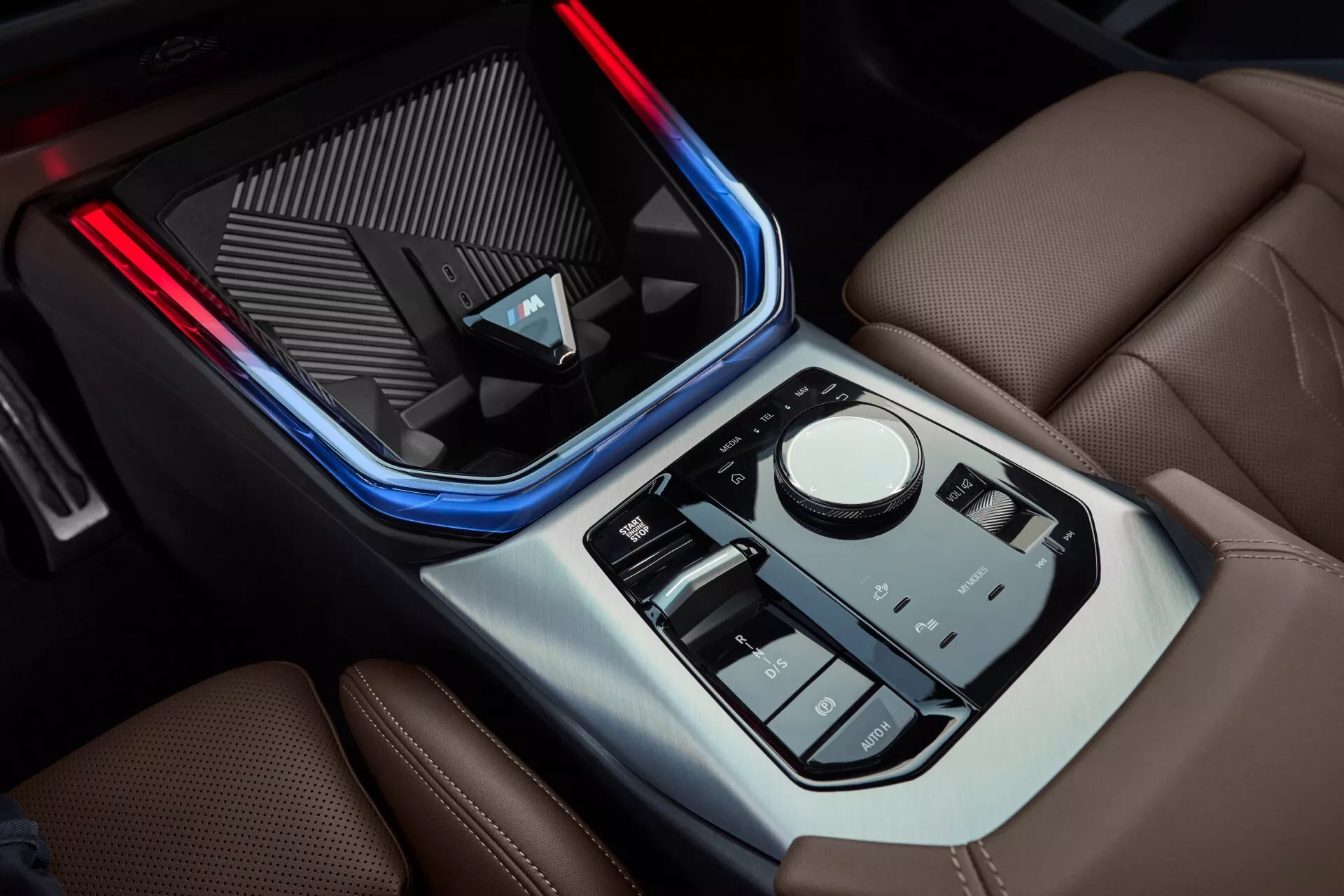 Zbliżenie konsoli środkowej BMW X3 2024 (G45) z przedstawieniem oświetlenia ambientowego, pokręteł sterujących, selektora biegów, schowka, uchwytu na kubek i materiałów przypominających skórę, ilustrujące nowoczesny design pojazdu, funkcje technologiczne oraz luksusowe wnętrze.