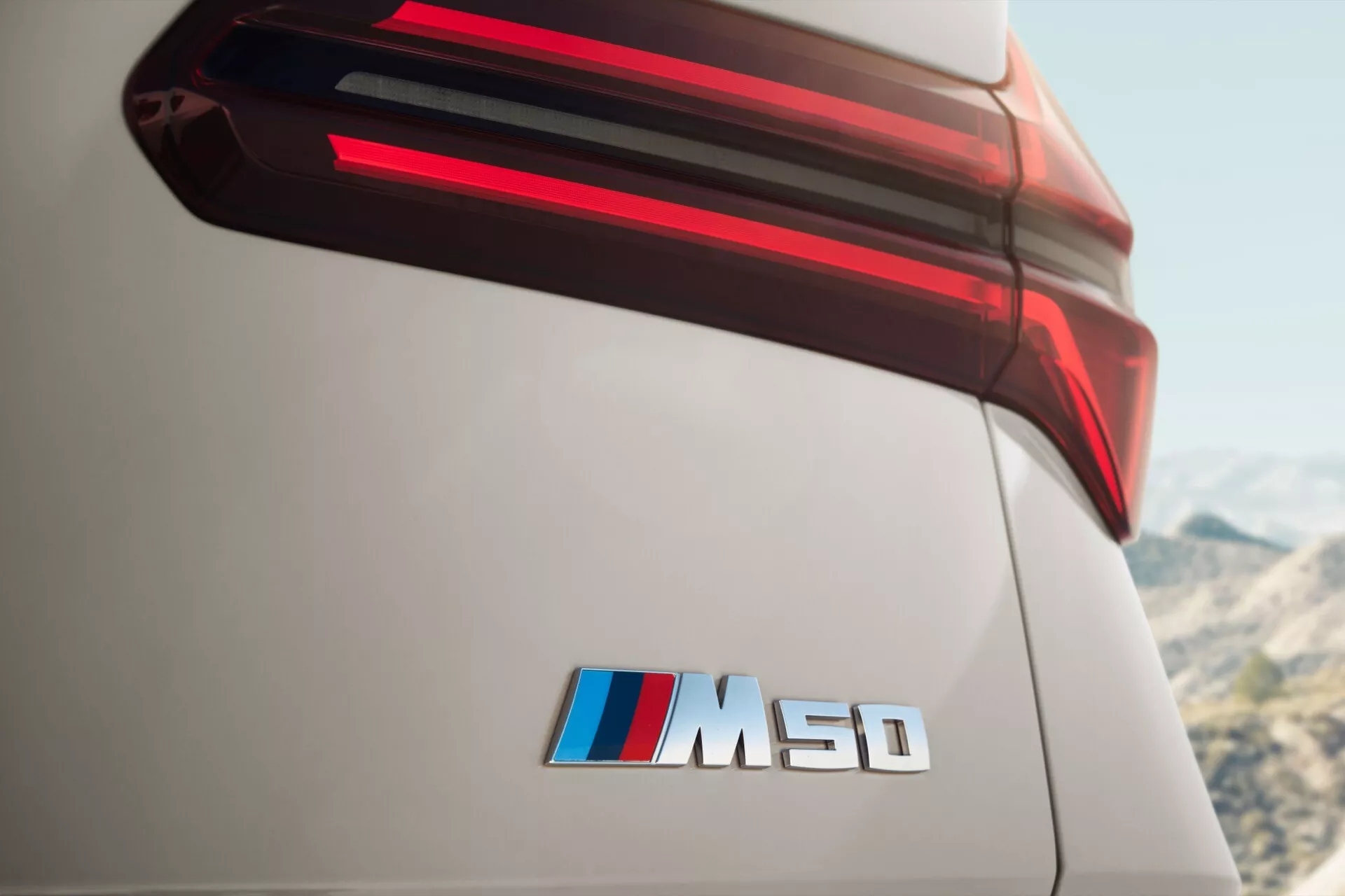 Zbliżenie tylnej części BMW X3 2024 (G45), podkreślające jego tylne światło LED i odznakę 'M50d' z kolorami M Performance. Perspektywa z niskiego kąta podkreśla premiumowy design samochodu i zaawansowaną technologię oświetlenia, z ostrą ostrością na tylne elementy na tle rozmytego zewnętrznego tła, sugerującego wydajność i luksus.