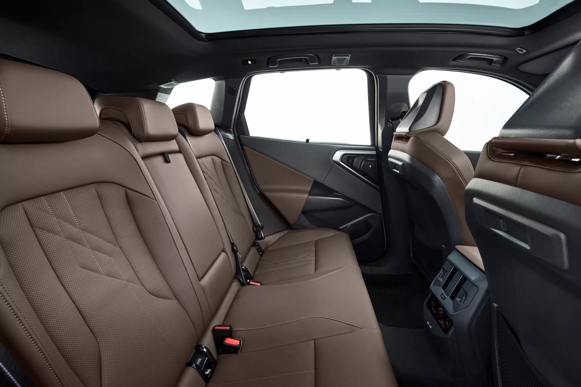 Widok wnętrza BMW X3 2024 (G45), prezentujący tylną część kabiny od strony pasażera. Na zdjęciu widać tylne siedzenia z brązowej skóry z luksusowymi przeszyciami, regulowane zagłówki, widoczne pasy bezpieczeństwa z czerwonymi przyciskami zwalniającymi, panel drzwi kierowcy z funkcjami sterowania i kratką głośnika, składany podłokietnik środkowy z uchwytami na kubki, duży panoramiczny dach, oświetlenie wnętrza, tylne nawiewy klimatyzacji na tylnej części konsoli centralnej i wykładzinę podłogową.
