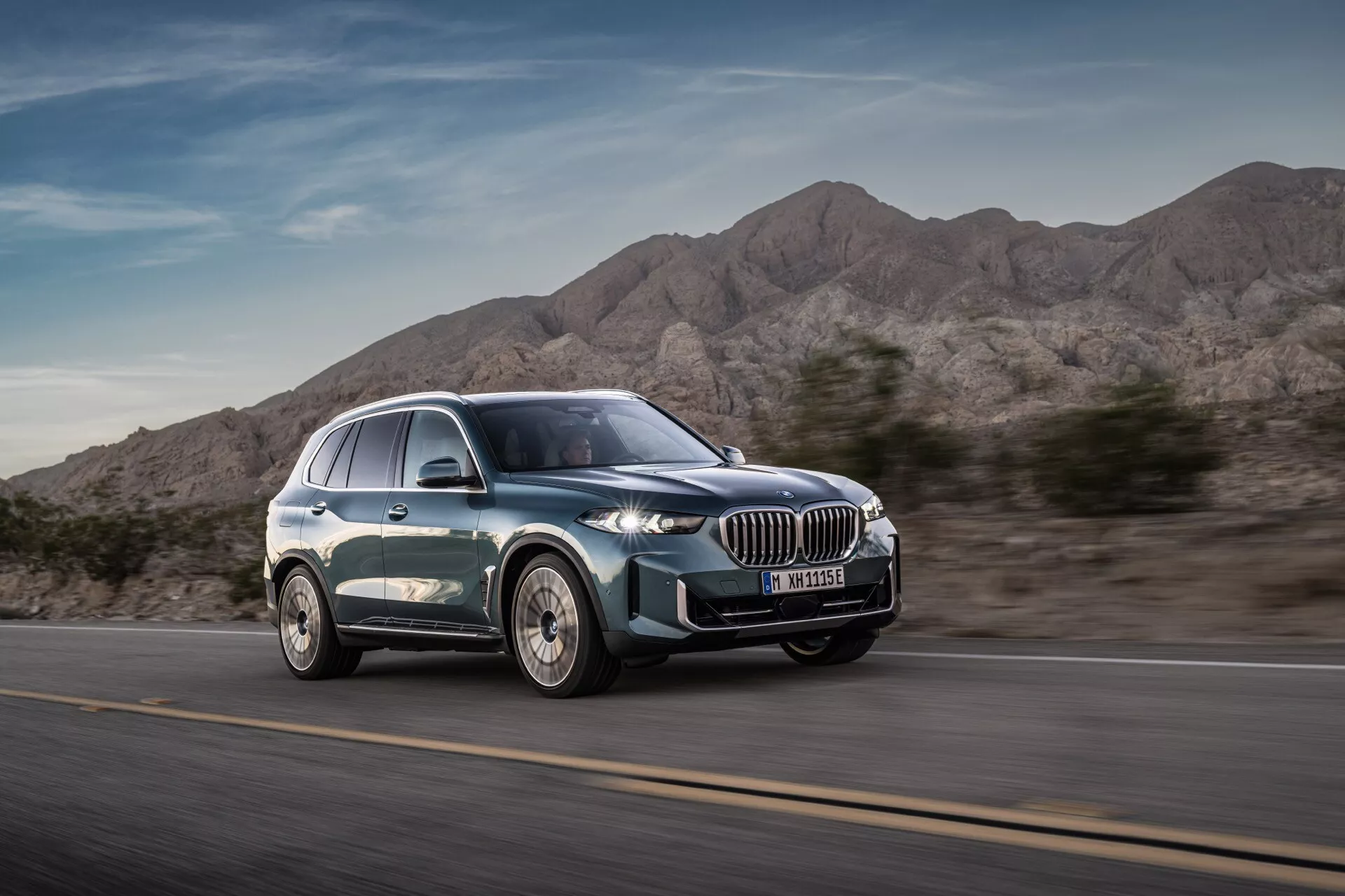 Nowoczesny luksusowy SUV BMW X5 2023 (G05-F95 Facelift) w ruchu z widoku trzy czwarte frontowe podkreślające dynamiczny design, duży grill nerkowy, wąskie reflektory LED, felgi aluminiowe wieloramienne i gładką sylwetkę z metalicznym lakierem