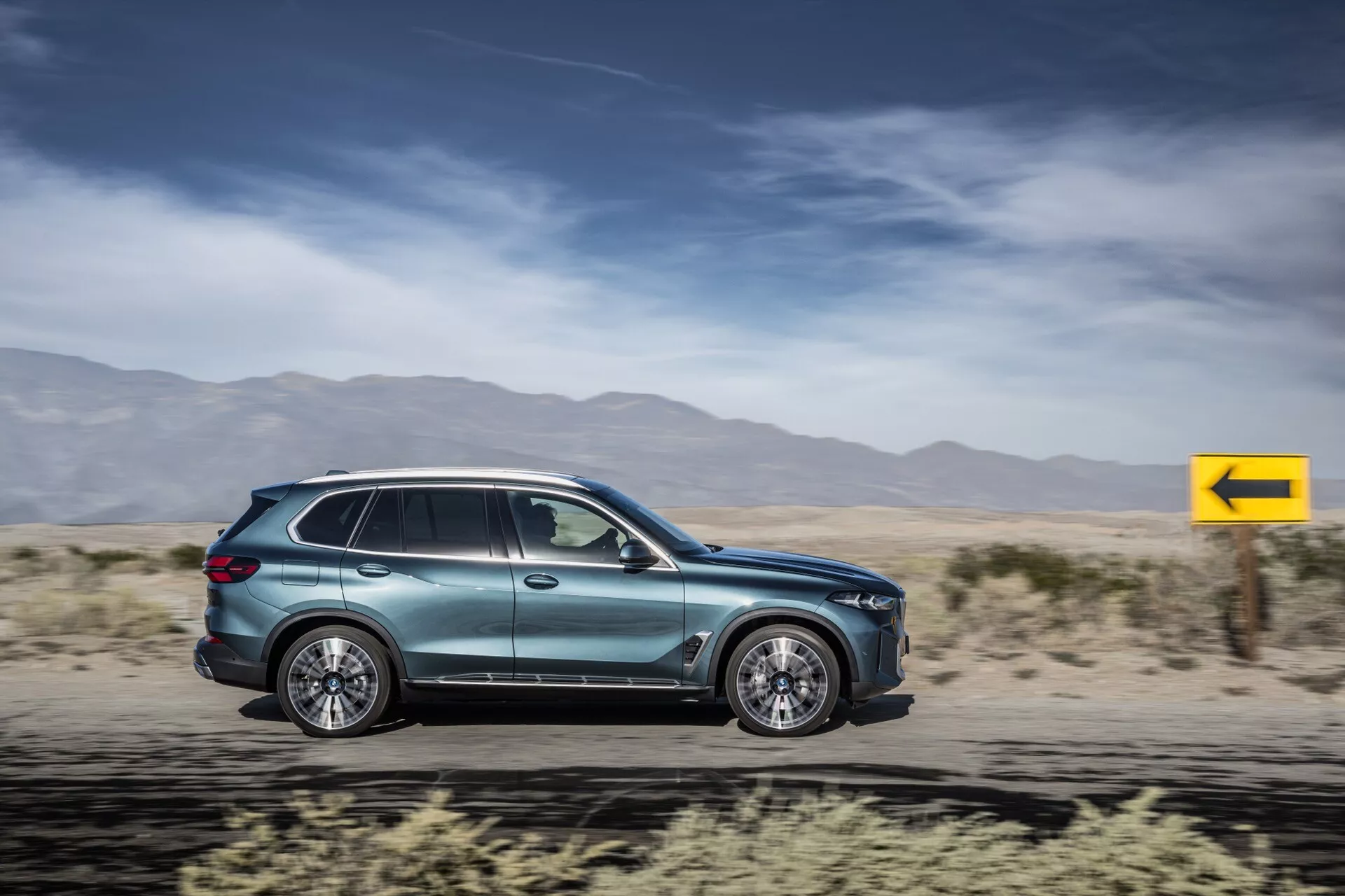 SUV BMW X5 2023 (G05-F95-Restyling) w ruchu, prezentujący aerodynamikę, felgi aluminiowe wieloramienne, zintegrowane klamki drzwi, aerodynamiczne lusterka boczne, przyciemniane szyby, boczny kierunkowskaz, metaliczny lakier oraz nowoczesny design tylnych świateł na rozmytym tle pustyni.