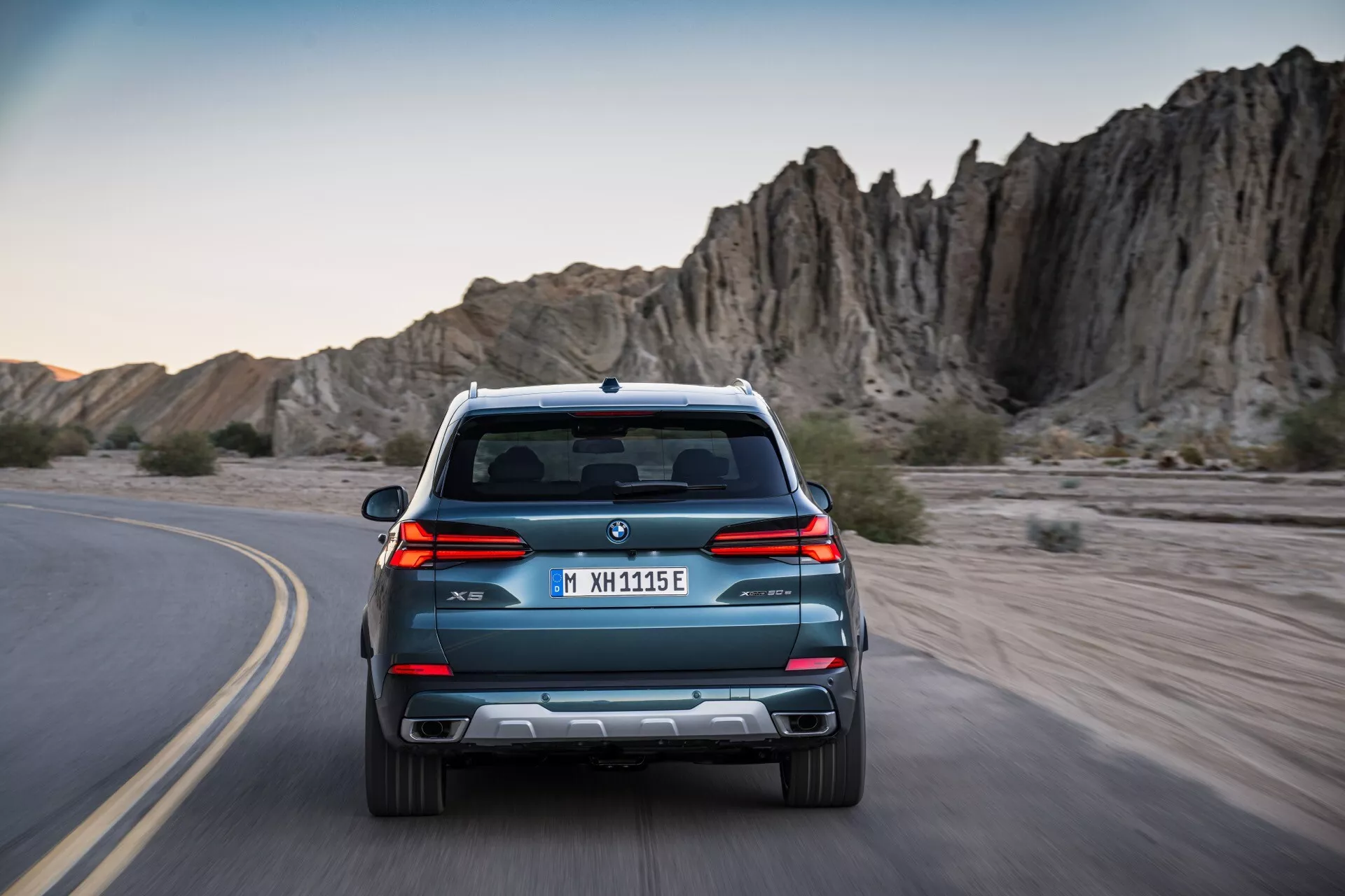 Tylny widok BMW X5 G05 F95 facelift 2023, jadącego po krętej drodze, z eleganckimi liniami, podwójnym wydechem i czujnikami parkowania, na tle przygodowego skalistego terenu