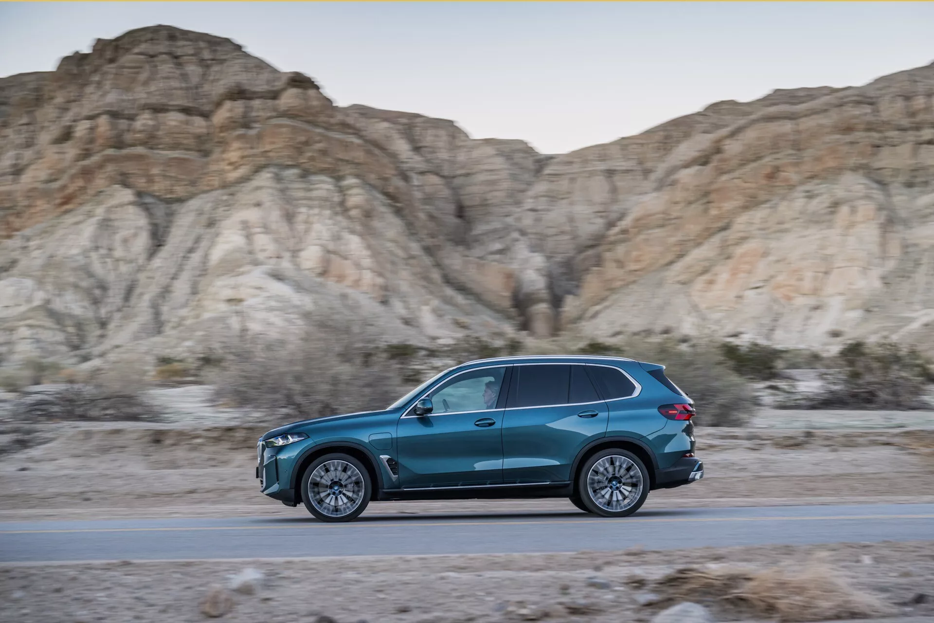 Dynamiczny boczny profil BMW X5 2023 (G05-F95 lifting) w ruchu, niebieskie SUV o kształcie łzy na drodze z tłem jałowych, skalistych wzgórz podczas złotej godziny, prezentujący elegancki design, wydajność aerodynamiczną, wieloramienną konstrukcję kół ze stopów i oświetlenie LED
