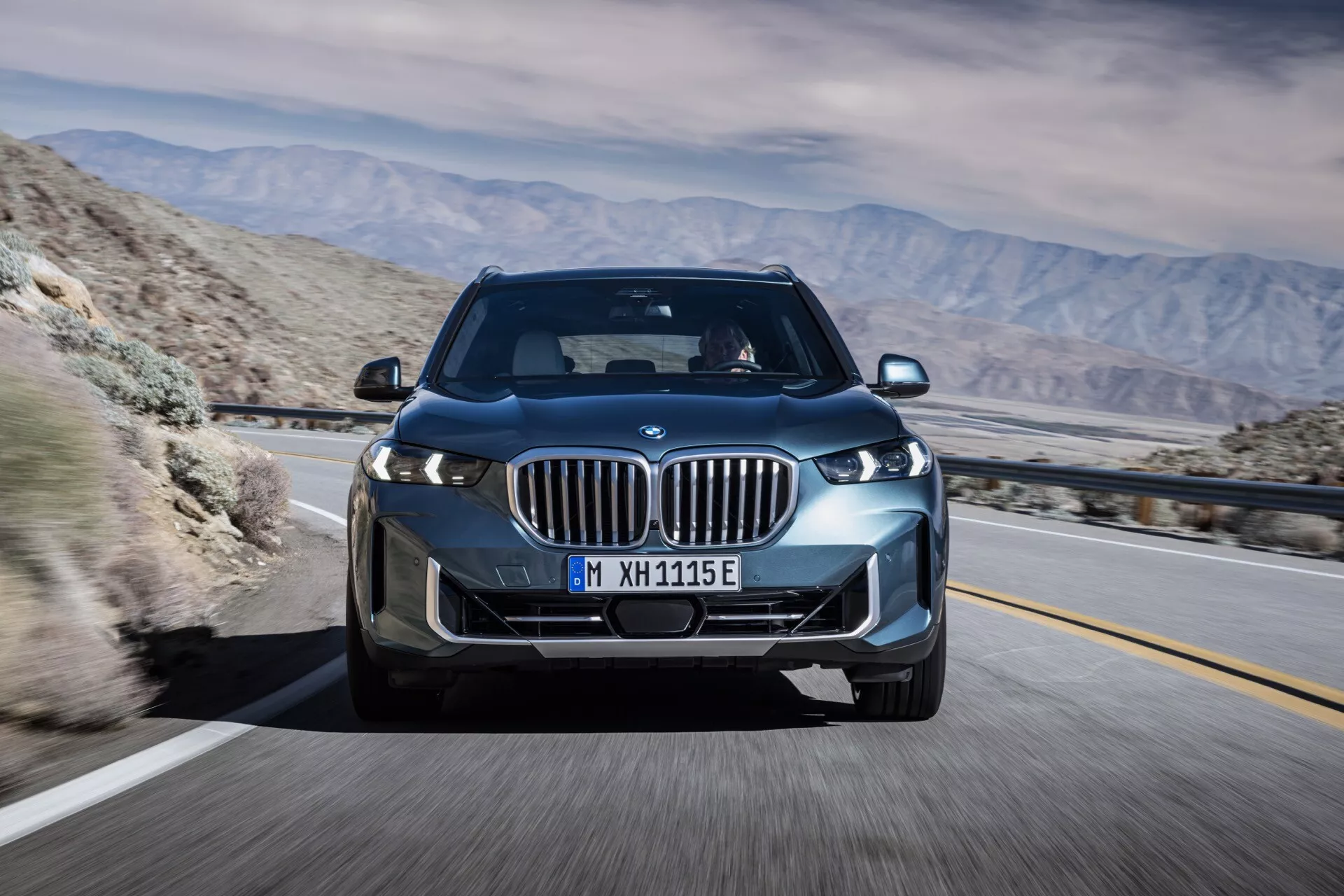 BMW X5 2023 (G05 F95 lifting) w ruchu, ujęte pod kątem przednim trzy czwarte, podkreślające jego diody LED, grill nerkowy z aktywnymi żaluzjami, felgi aluminiowe i eleganckie aerodynamiczne wzornictwo na tle górskiej pustyni, sugerując zaawansowaną technologię i potencjalne możliwości elektryczne lub hybrydowe