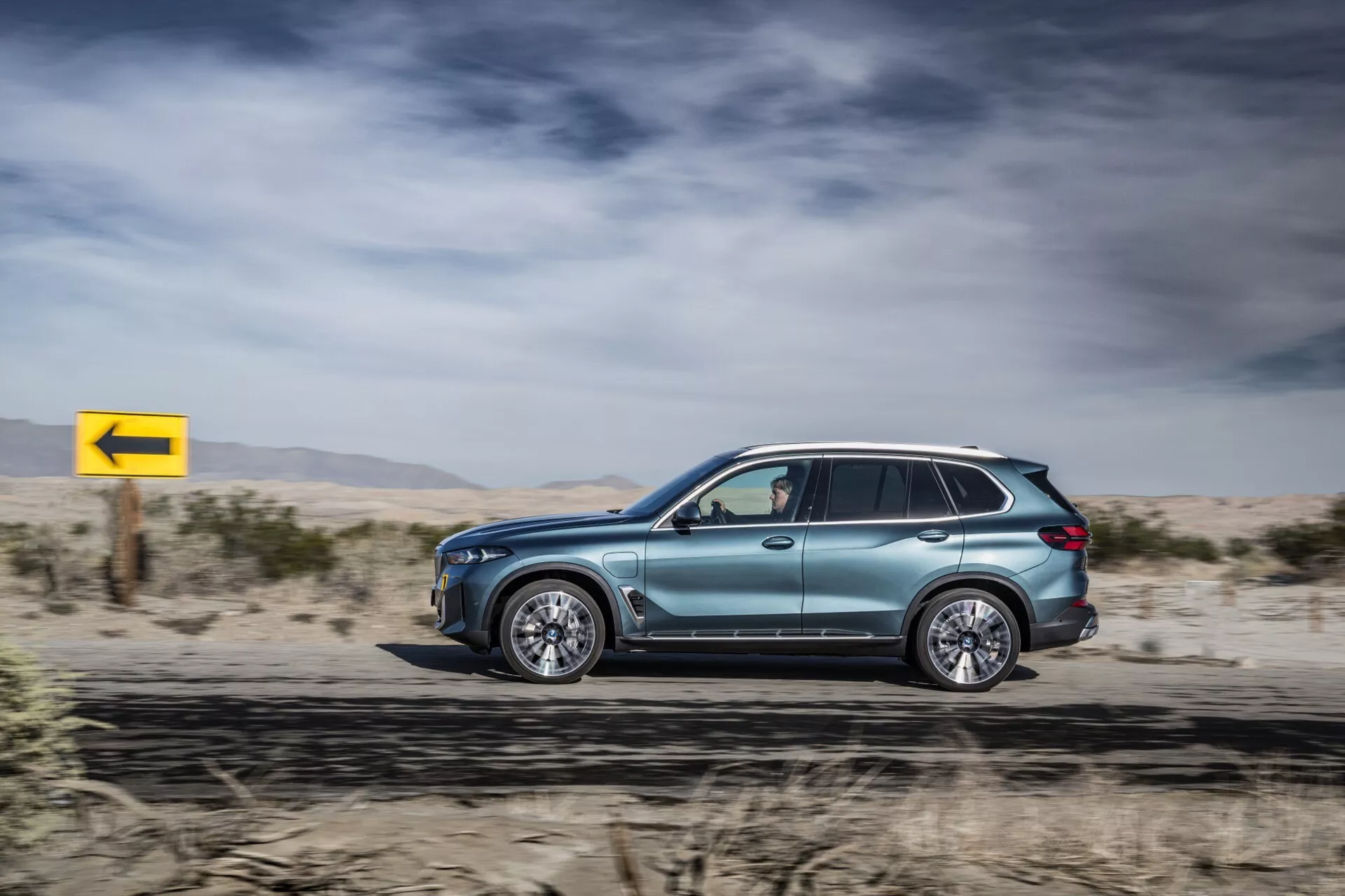 Boczny profil w ruchu BMW X5 2023 (G05-F95 Facelift), prezentujący metaliczny lakier, aerodynamiczny design, luksusowe koła ze stopu, przyciemniane szyby z lusterkami z kierunkowskazami, relingi dachowe i zaawansowaną technologię LED, na tle pustynnym z dynamicznym żółtym znakiem skrętu w lewo.