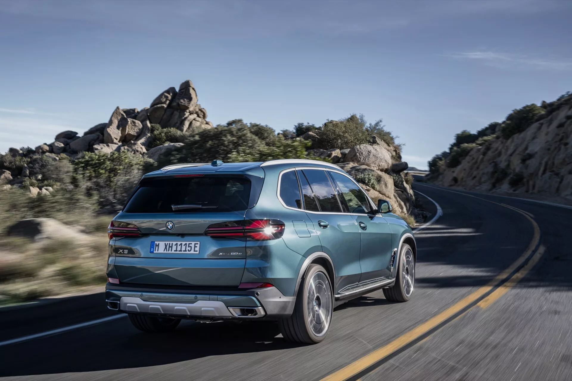 BMW X5 2023 (lifting G05-F95) w ruchu, uchwycone z tylnego widoku trzy czwarte podkreślające sportowy design SUV, nowoczesną klapę bagażnika ze smukłymi światłami tylnymi, dwukolorowe koła z stopu i dowody na elektryfikację na krętej drodze w jasnym świetle dziennym