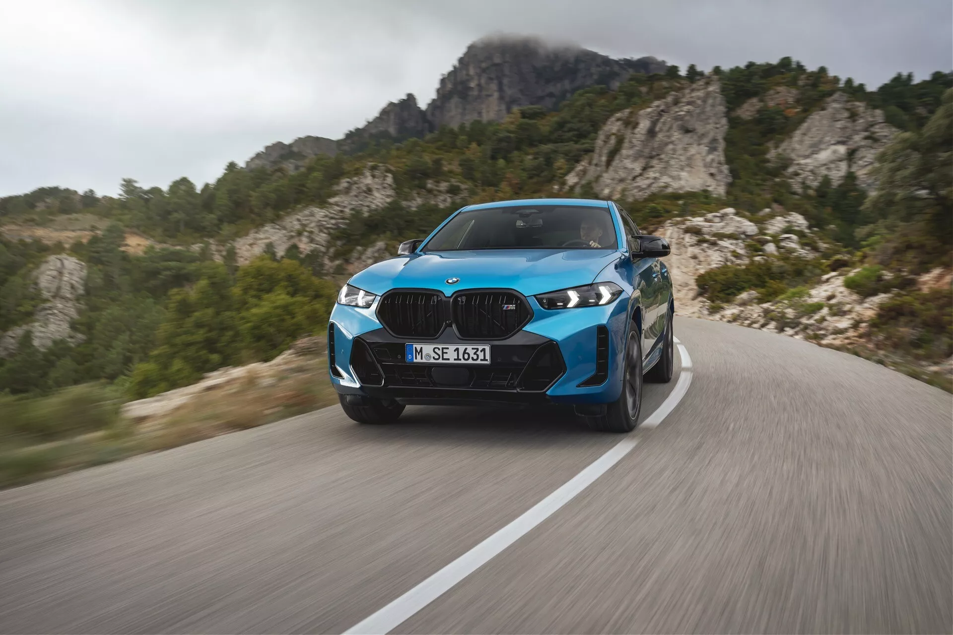 BMW X6 2023 (G06-F96-facelift) w żywym niebieskim kolorze, prezentujące nowoczesne luksusowe wykonanie na krętej górskiej drodze z dynamicznym rozmyciem ruchu, zaawansowanymi reflektorami LED, eleganckimi elementami projektu i sportowymi kołami, na tle skalistego terenu górskiego.