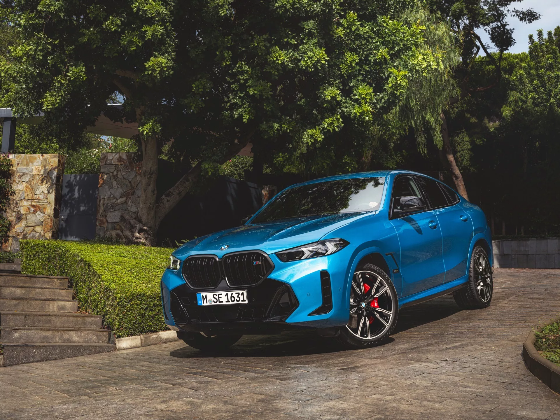 BMW X6 2023 (G06-F96-Facelift) w kolorze niebieskim, zaparkowane pod kątem ukazujące przednią część, nerkowatą atrapę, reflektory LED lub laserowe, sportowe felgi aluminiowe z czerwonymi zaciskami hamulcowymi, konturowane linie maski i aerodynamiczne lusterka boczne z wbudowanymi kierunkowskazami, uchwycone w naturalnym świetle dzieńnym z roślinnością i architektonicznymi schodami w tle
