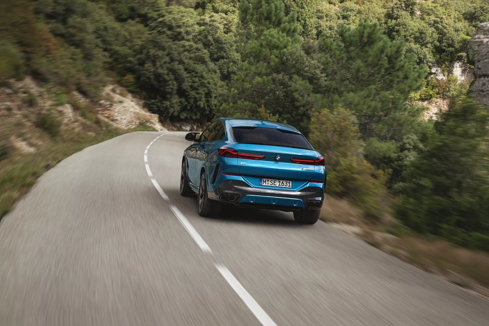 Dynamiczny widok z tyłu niebieskiego BMW X6 2023 (G06-F96-Facelift) z sportowym designem i nowoczesnymi lampami tylnymi na krętej drodze pośród naturalnego krajobrazu