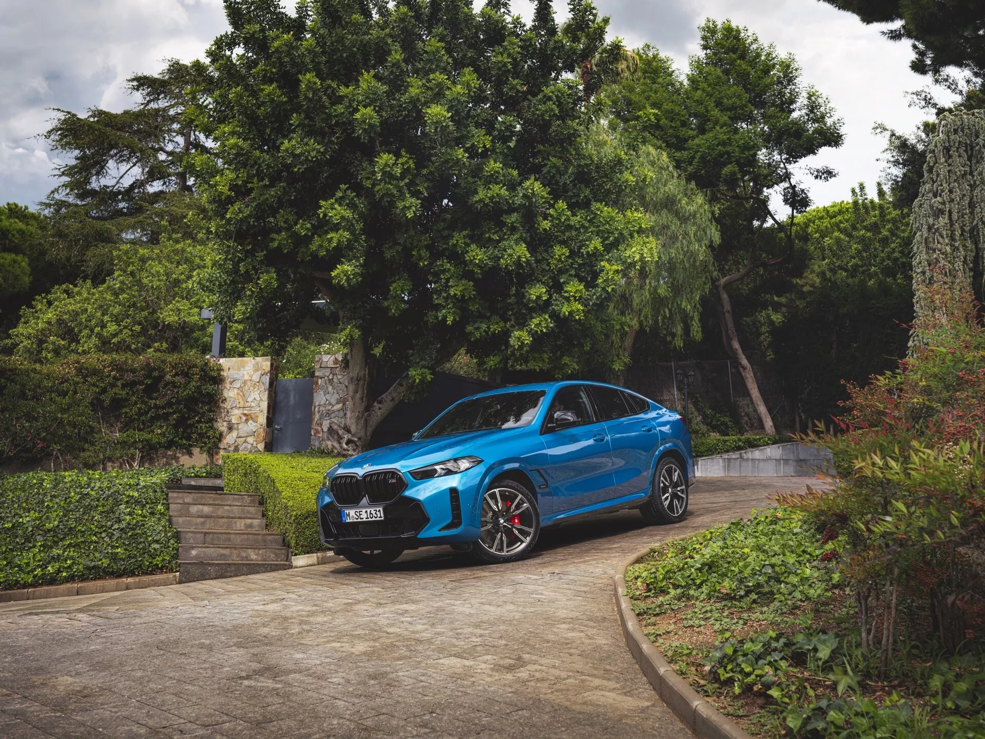 BMW X6 2023 (G06-F96-Facelifting) w kolorze niebieskim, widok z przodu pod trzech czwartych, zaparkowane na podjeździe otoczonym zielenią, pokazujące przedni zderzak, grill, maskę, przednią szybę, profil kierowcy, reflektory LED, sportowe koła z czerwonymi zaciskami hamulcowymi i aerodynamiczny design
