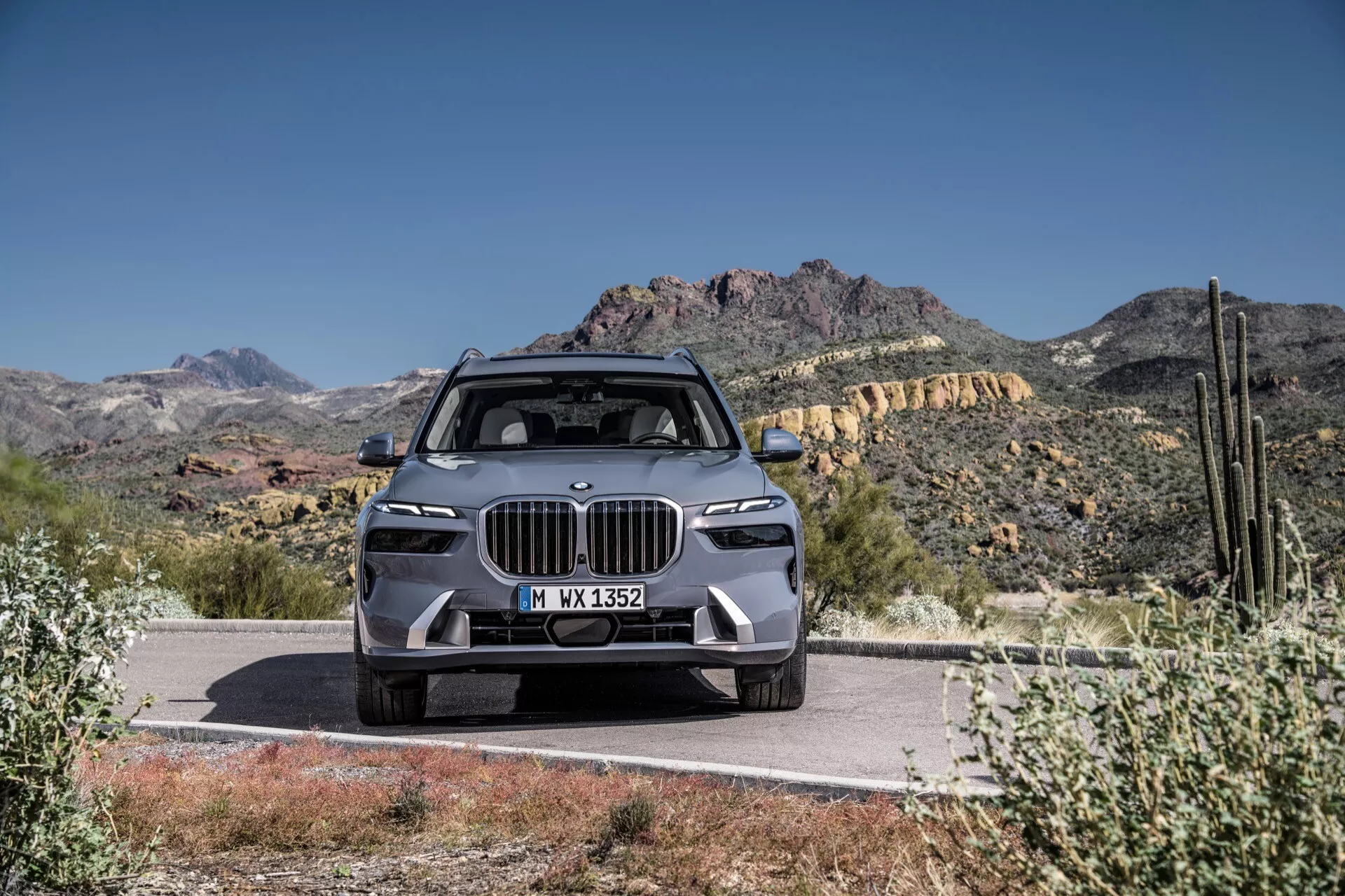 Luksusowy SUV BMW X7 2022 (G07-Facelift), prezentujący nowoczesny design z dużą atrapą w stylu nerkowym, wąskimi reflektorami LED, zaawansowanym zderzakiem przednim z funkcjami ADAS, wyrzeźbioną maską i felgami z lekkich stopów, górujący na drodze na tle pustynnych gór pod jasnoniebieskim niebem