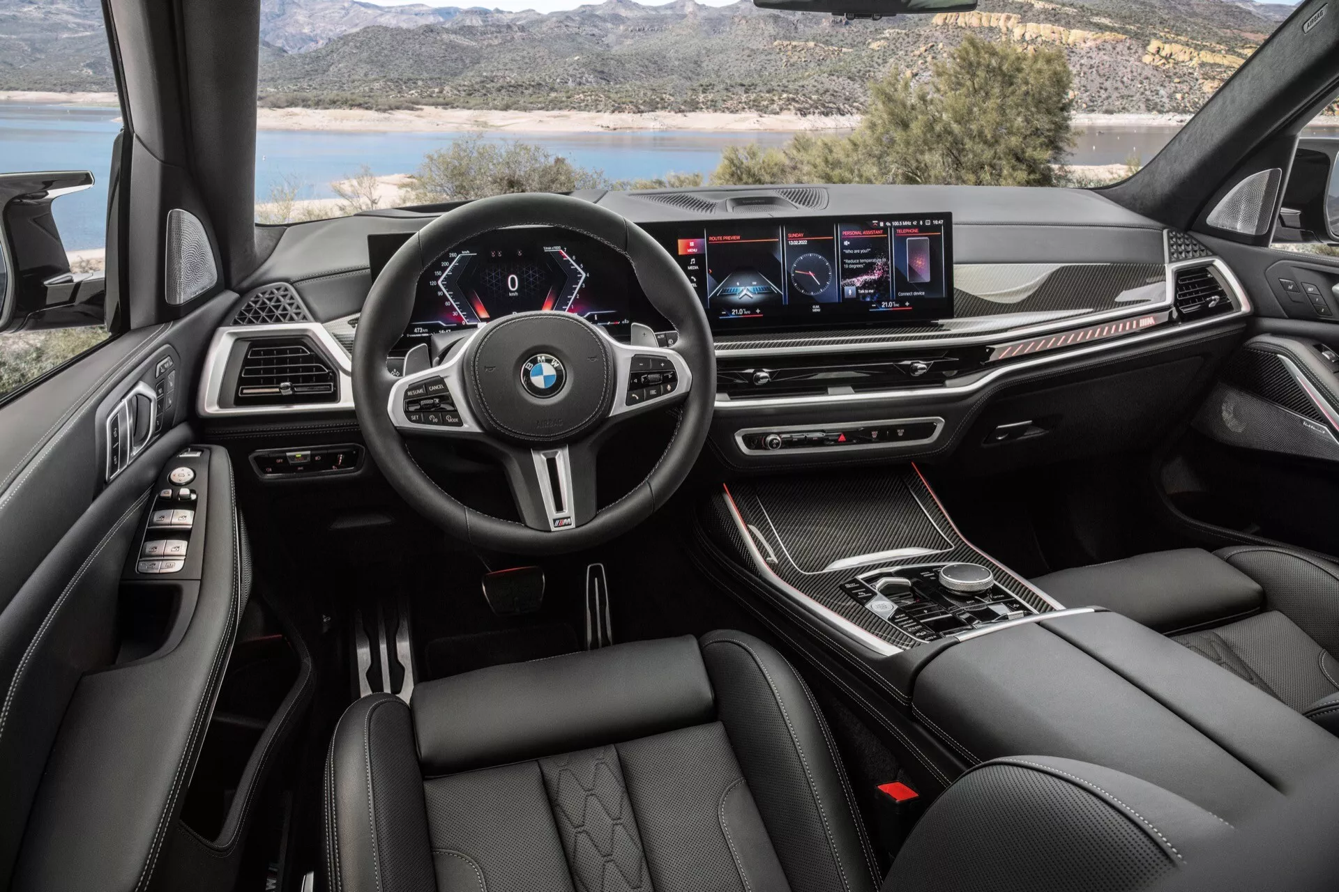 Widok wnętrza BMW X7 2022 (G07 Facelift) z perspektywy kierowcy, przedstawiający wielofunkcyjną kierownicę z logiem BMW, cyfrowy zestaw wskaźników, szerokoekranowy wyświetlacz systemu infotainment, przyciski kontroli klimatyzacji, selektor automatycznej skrzyni biegów, luksusowe materiały, sportowe i wygodne fotele oraz oświetlenie ambientowe