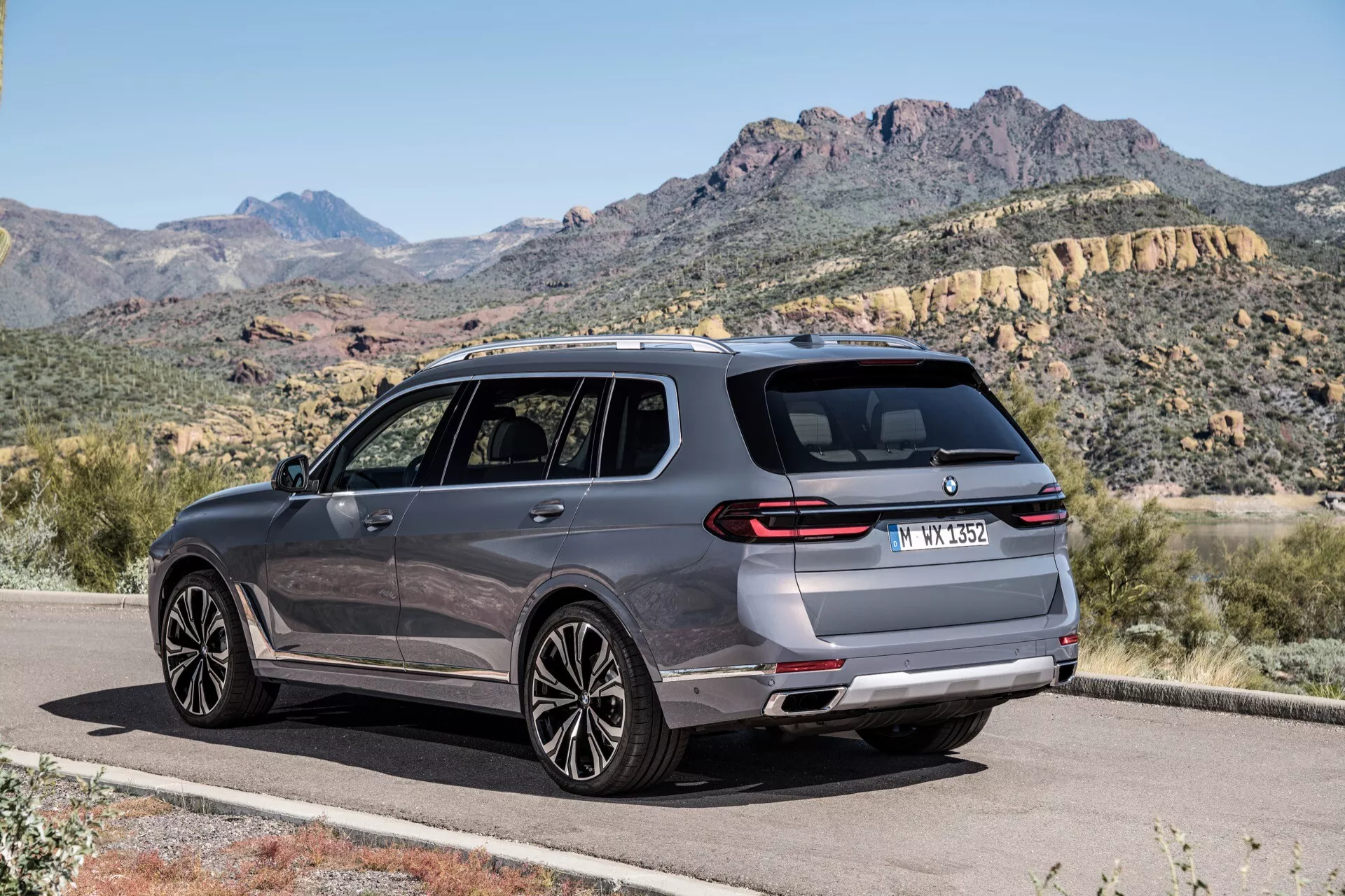 Nowoczesne SUV BMW X7 2022 (G07 Facelift) w metalicznym lakierze na utwardzonej drodze przeciwko górskiemu krajobrazowi, prezentujące tylny i boczny profil z trzech czwartych, z widocznymi tylnymi światłami, tylnym zderzakiem, czujnikami parkowania, podwójnymi rurami wydechowymi, felgami ze stopu o wielu szprychach, oknami z chromowanymi obramowaniami, lusterkami bocznymi z wbudowanymi kierunkowskazami i relingami dachowymi