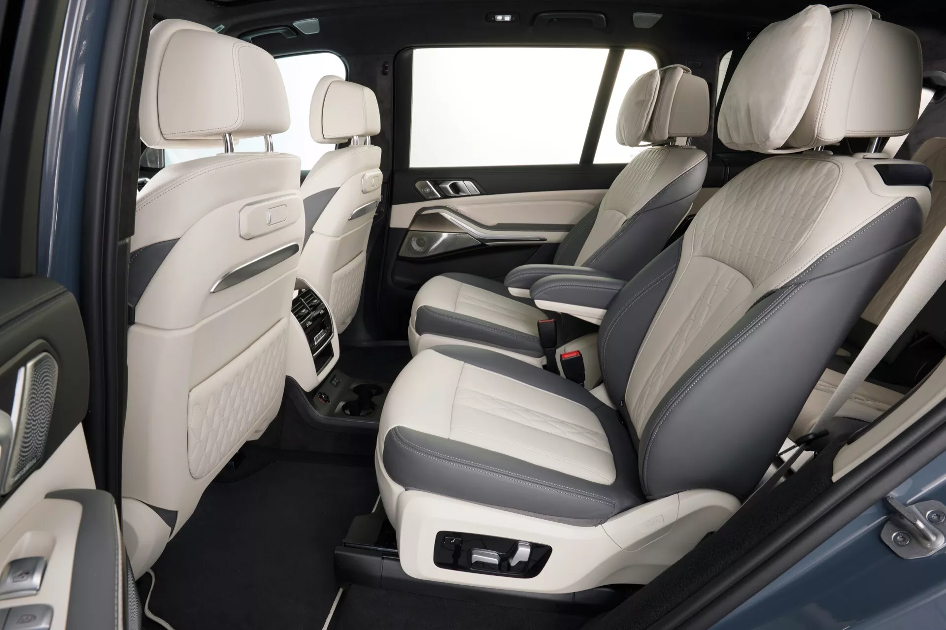 Widok wnętrza BMW X7 (G07 Facelift) z 2022 roku z akcentem na tylny przedział pasażerski, z tapicerką z dwukolorowej skóry, zaawansowanymi kontrolami systemu audio na panelach drzwi, konsolą środkową z akcentami z materiałów wysokiej jakości, wykładziną dywanową, obszerną przestrzenią na nogi i jasnym schematem kolorystycznym wnętrza dla przestronnego wrażenia kabiny