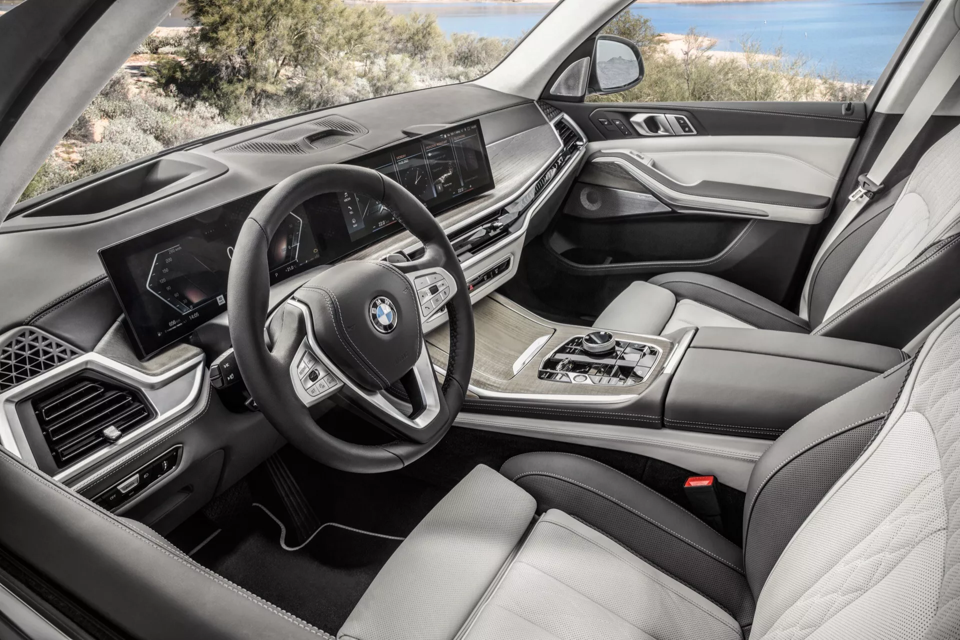 Widok wnętrza BMW X7 z 2022 roku (G07-Facelift) od strony pasażera, przedstawiający deskę rozdzielczą z ekranem systemu infotainment, wielofunkcyjną kierownicę, automatyczną dźwignię zmiany biegów, pokrętło kontrolne multimediów iDrive, klimatyzację dwustrefową, luksusowe skórzane siedzenia, drewniane i metaliczne akcenty oraz cyfrowy kokpit.