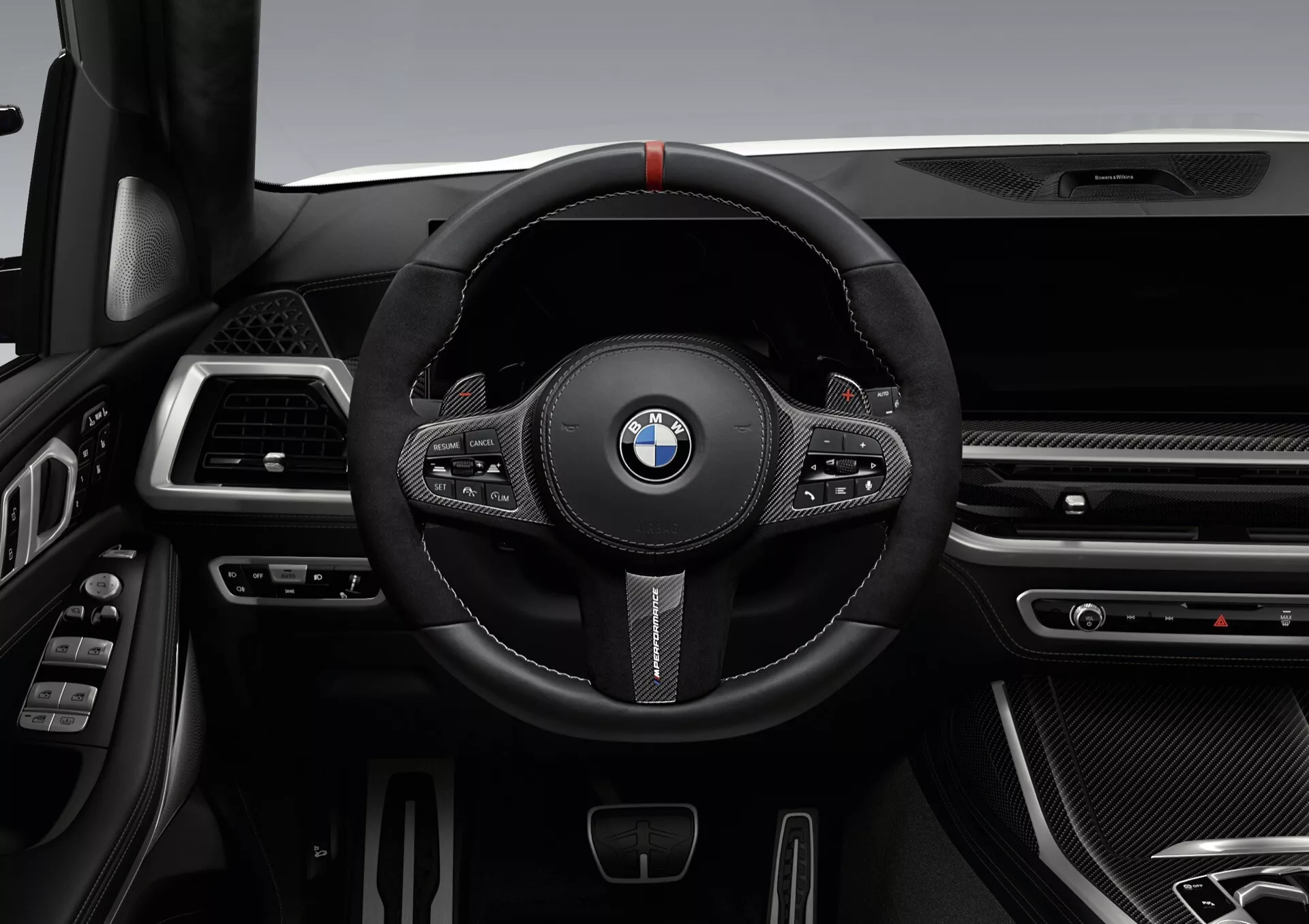 Wnętrze BMW X7 2022 (G07-Facelift) skupiające się na sportowej kierownicy pokrytej czarną skórą z czerwonym znacznikiem, przyciskami wielofunkcyjnymi i łopatkami zmiany biegów, przeciwko zaawansowanej desce rozdzielczej ze smukłymi nawiewami, teksturami przypominającymi włókno węglowe i chromowanymi akcentami
