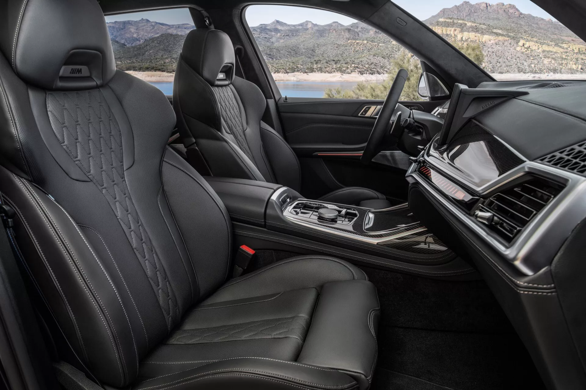 Widok wnętrza BMW X7 2022 (G07-Facelift), skupiający się na luksusowym przednim wnętrzu z czarnymi skórzanymi siedzeniami z bocznymi wzmocnieniami i wytłaczanymi zagłówkami, elegancka deska rozdzielcza z ekranem systemu informacyjno-rozrywkowego, centralna konsola z ergonomiczną dźwignią zmiany biegów i wykończenia wysokiej jakości wskazujące na pojazd luksusowy.