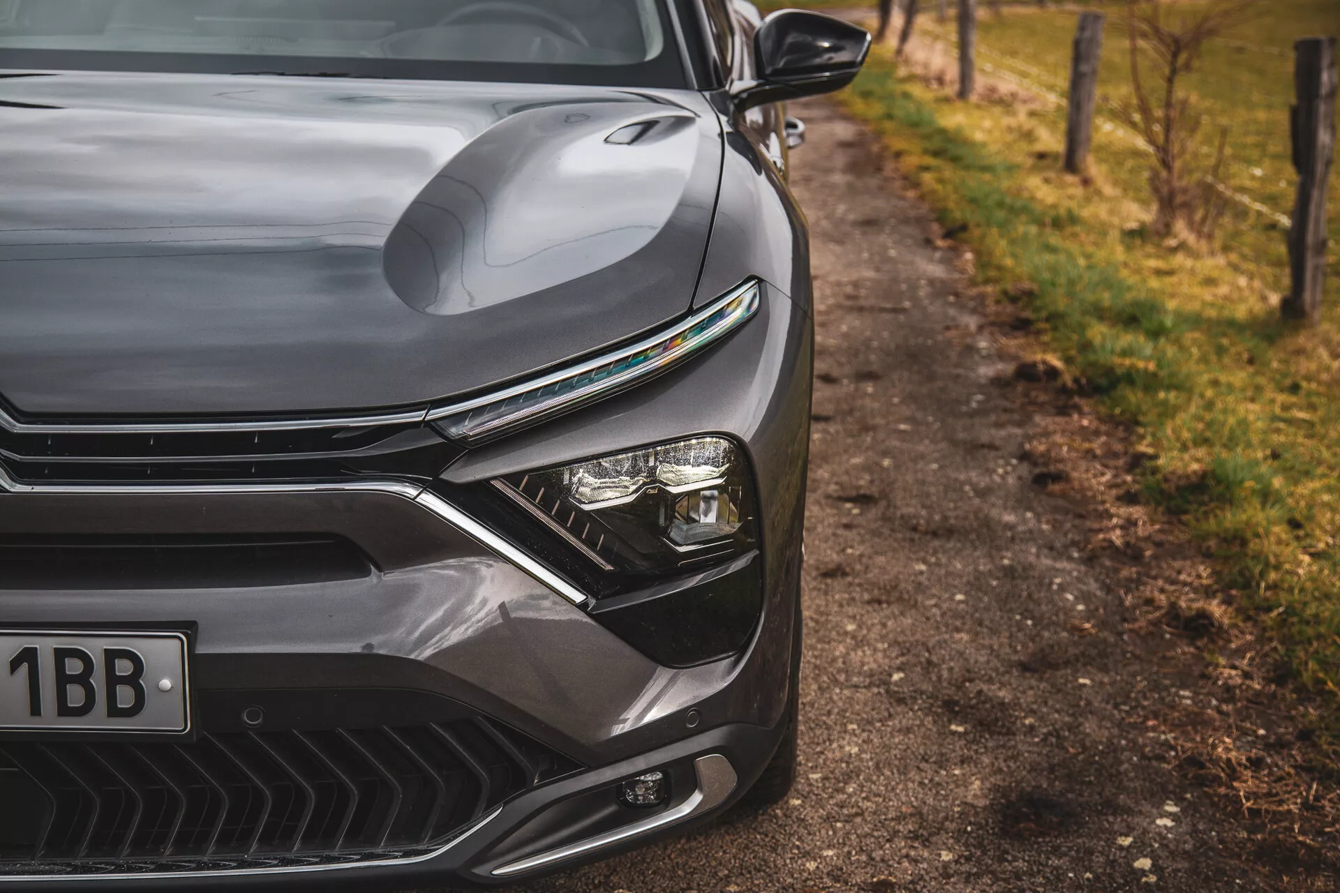 Zbliżenie na Citroena C5-X 2022 ukazujące elegancki design przedniej lewej strony, w tym konturowaną maskę, wyrazistą osłonę chłodnicy z poziomymi liniami, zaawansowany system reflektorów LED i aerodynamicznie ukształtowany przedni zderzak z otworami na wloty powietrza, na nieostrym naturalnym tle przy drodze.
