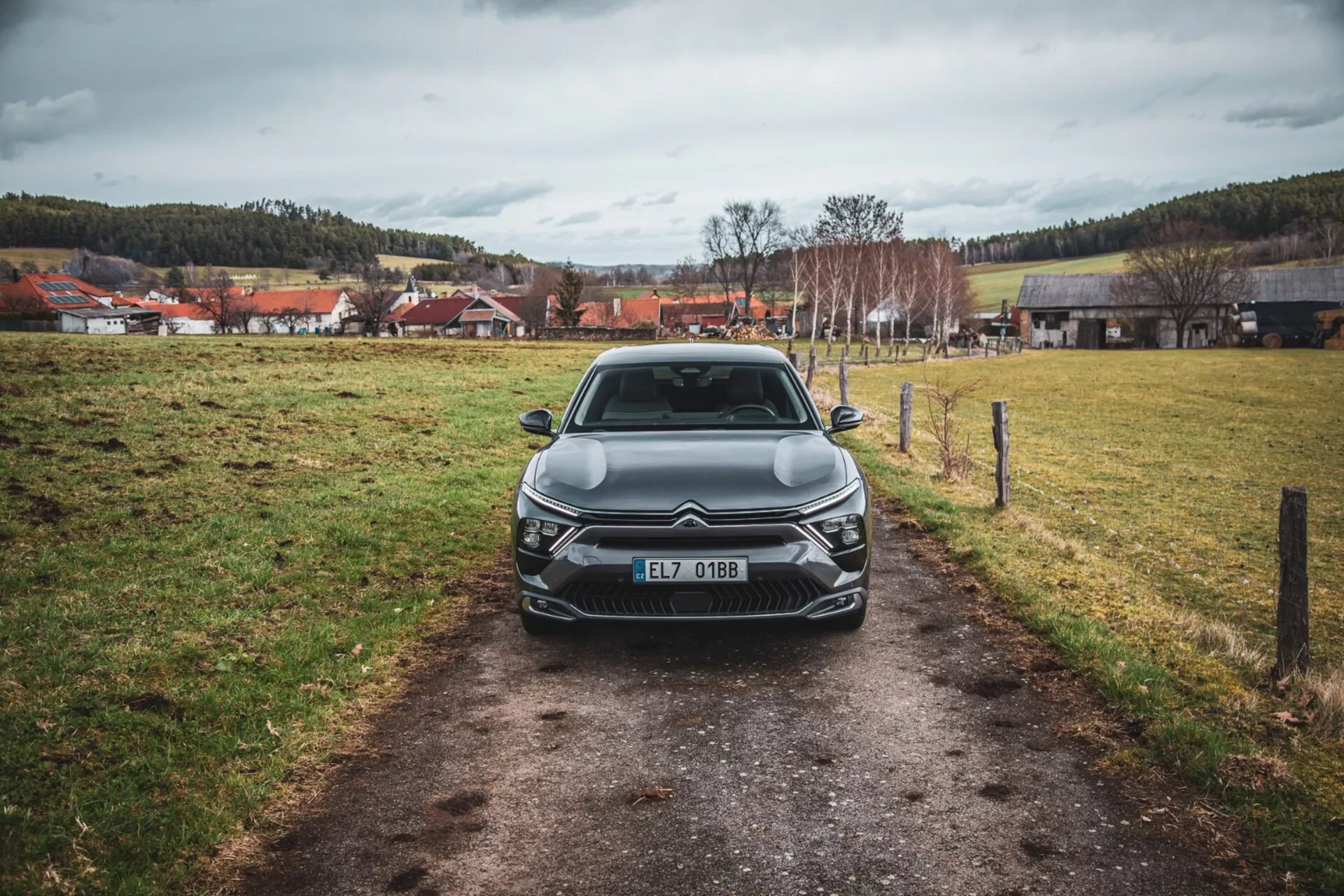 Widok frontalny Citroëna C5-X 2022 na wąskiej ścieżce na trawiastym polu pod zachmurzonym niebem, prezentujący jego elegancki design, złożoną kratę z emblematem, kątowe jednostki świateł LED, rzeźbione linie maski i dodatkowe elementy oświetlenia przedniego, zarejestrowany w Czechach.