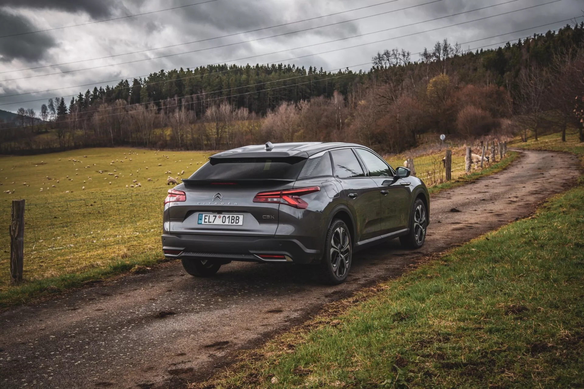 Nowoczesny hatchback Citroen C5-X 2022 zaparkowany na nieutwardzonej wiejskiej drodze, widok z tyłu i boku ukazujący aerodynamiczny design z spoilerem i anteną w kształcie płetwy rekina, smukłe poziome tylne światła i duże koła o wieloramiennej konstrukcji, sugerujące wydajność i luksus poza miejskim otoczeniem.