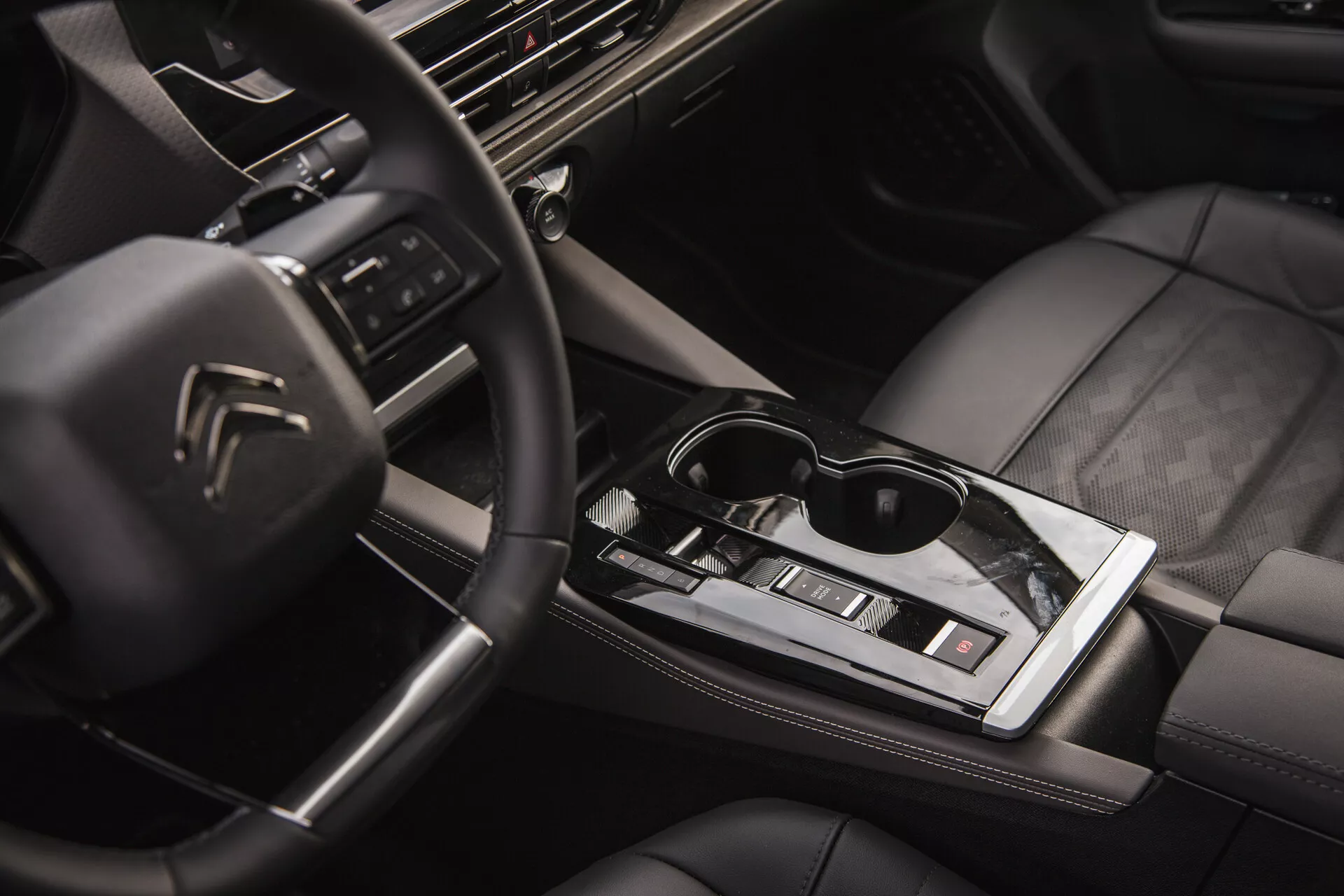 Widok wnętrza Citroën C5-X 2022 z wielofunkcyjną kierownicą z logiem, zaawansowaną technologicznie deską rozdzielczą z systemem infotainment, eleganckim automatycznym dźwignią zmiany biegów, kontrolą klimatyzacji, siedzeniami tapicerowanymi skórą z wysokiej jakości szwami i elementem stylu życia z okularami przeciwsłonecznymi na błyszczącej środkowej powierzchni