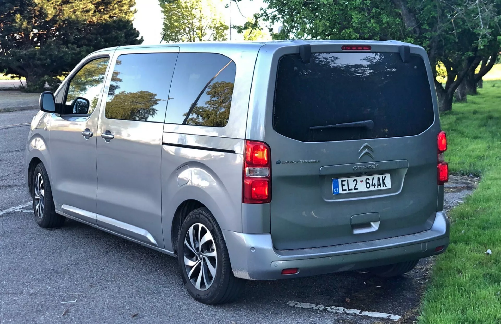 Citroën Spacetourer 2016 uchwycony pod trzykrawędziowym kątem z tyłu, podkreślający prawą tylną stronę z tylną klapą, odmrażaczem tylnej szyby, tylną wycieraczką, podwójnymi tylnymi światłami, centralnie zamontowaną tablicą rejestracyjną, przesuwanymi drzwiami bocznymi, tylnymi oknami sugerującymi możliwe dodatkowe siedzenia oraz srebrnymi, wieloramiennymi felgami z metalu; zaparkowany na ulicy na przedmieściach lub w otoczeniu przypominającym park z widocznymi drzewami i trawą.