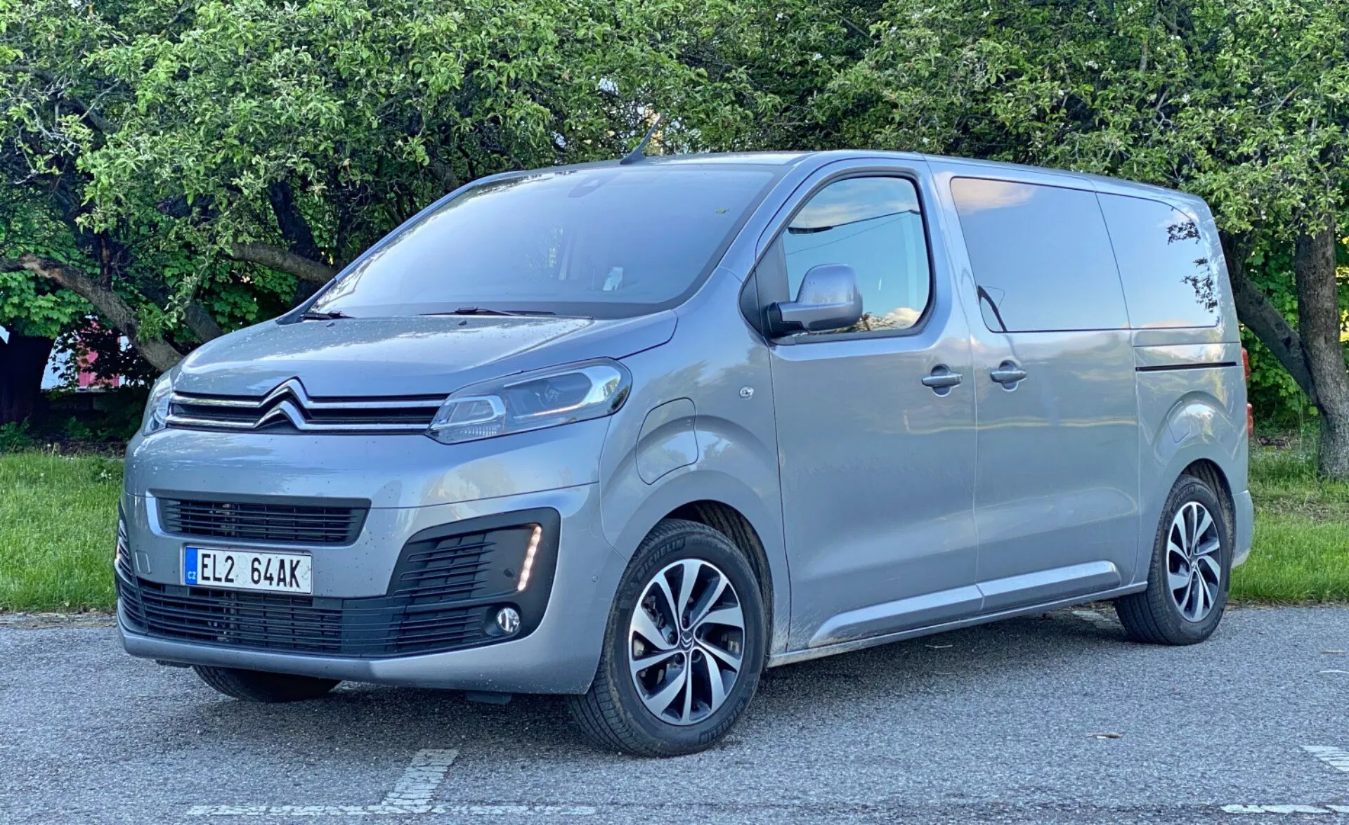Citroen Spacetourer 2016 na spokojnym terenie na świeżym powietrzu, widok z przodu trzy czwarte ukazujący przedni grill z charakterystycznym wzorem i logo marki, eleganckie reflektory LED, przedni zderzak z reflektorami przeciwmgielnymi i wlotami powietrza, przesuwane drzwi boczne z wbudowanym bocznym stopniem, okna pasażerskie za przednimi drzwiami, nowoczesne aluminiowe felgi z wieloszprychowym wzorem, metaliczny lakier, duże lusterka boczne z wbudowanymi kierunkowskazami, subtelne przetłoczenia wzdłuż bocznego panelu.