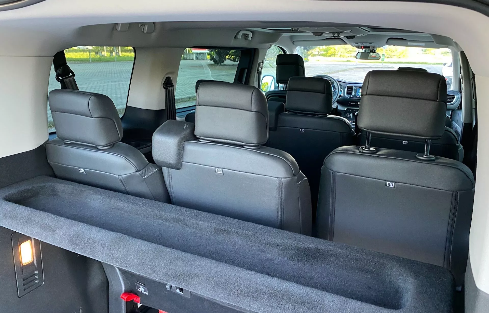 Widok wnętrza Citroëna Spacetourer 2016 z perspektywy tylnej, z przestronną kabiną z trzema rzędami siedzeń. Przestrzeń ładunkowa na pierwszym planie zawiera wykładzinę dywanową i pokrowiec ładunkowy z tkaniny. Trzeci rząd składa się z dwóch indywidualnych siedzeń z podniesionymi zagłówkami, podczas gdy drugi rząd ma trzy większe siedzenia z podniesionymi zagłówkami i pasami bezpieczeństwa. Częściowy widok na przednią kabinę odsłania deskę rozdzielczą, konsolę środkową i duży ekran informacyjno-rozrywkowy. Podsufitka ma wbudowane oświetlenie kabiny i uchwyty dla pasażerów, co podkreśla komfort i nowoczesne funkcje.