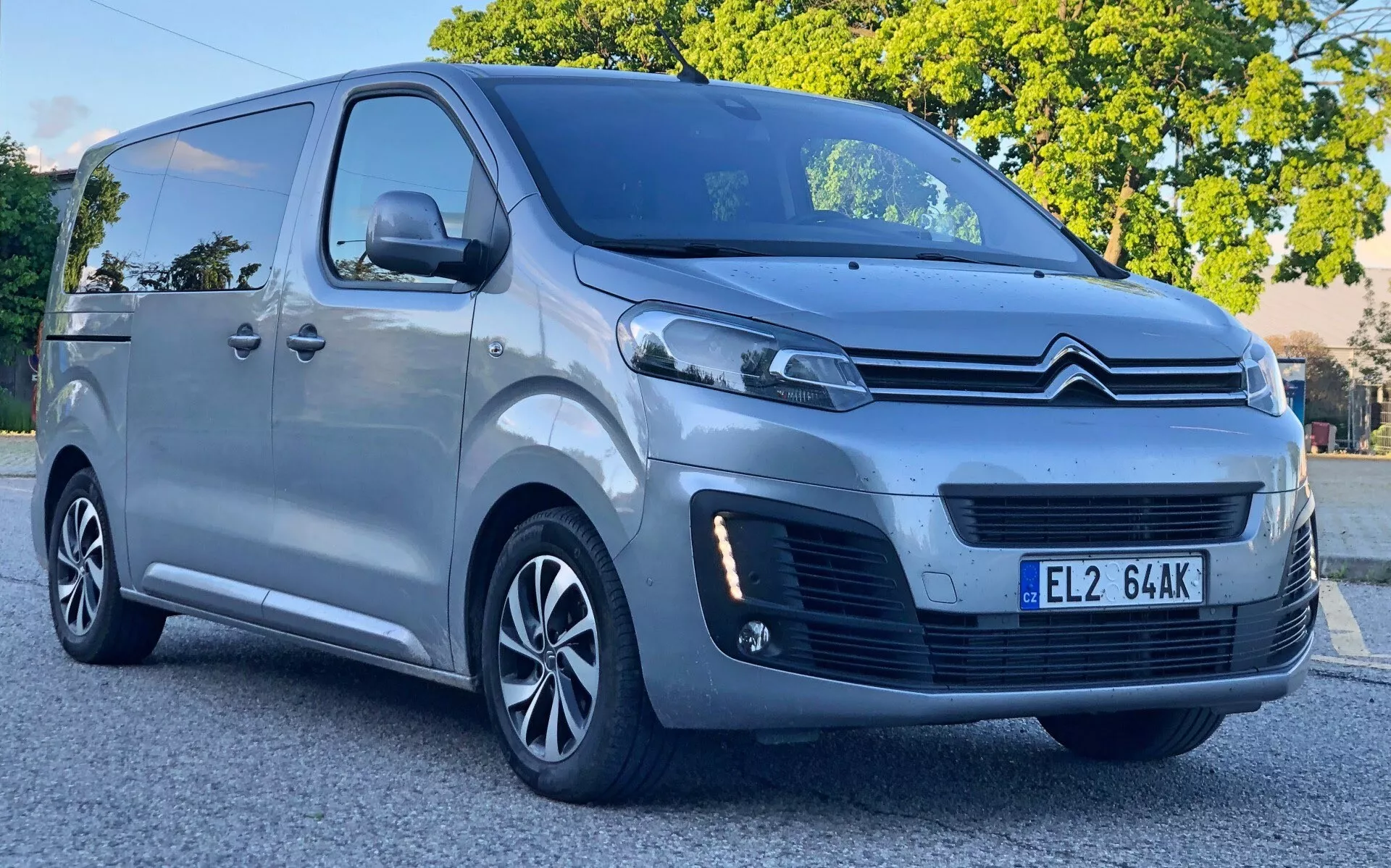 Obraz Citroëna Spacetourera 2016 widziany z perspektywy przedniego trójkąta, ukazujący przedni grill z chromowanym logo Citroëna z podwójnym szewronem, eleganckie reflektory prawdopodobnie z dziennymi światłami LED i światła przeciwmgielne poniżej. Van ma duże boczne okna, felgi aluminiowe i boczne drzwi przesuwne, co wskazuje na projekt zarówno dla pasażerów, jak i do lekkich zastosowań komercyjnych. Jest zaparkowany na ulicy w świetle dziennym, zapewniając wyraźny widok na jego cechy technologiczne i ogólny design.