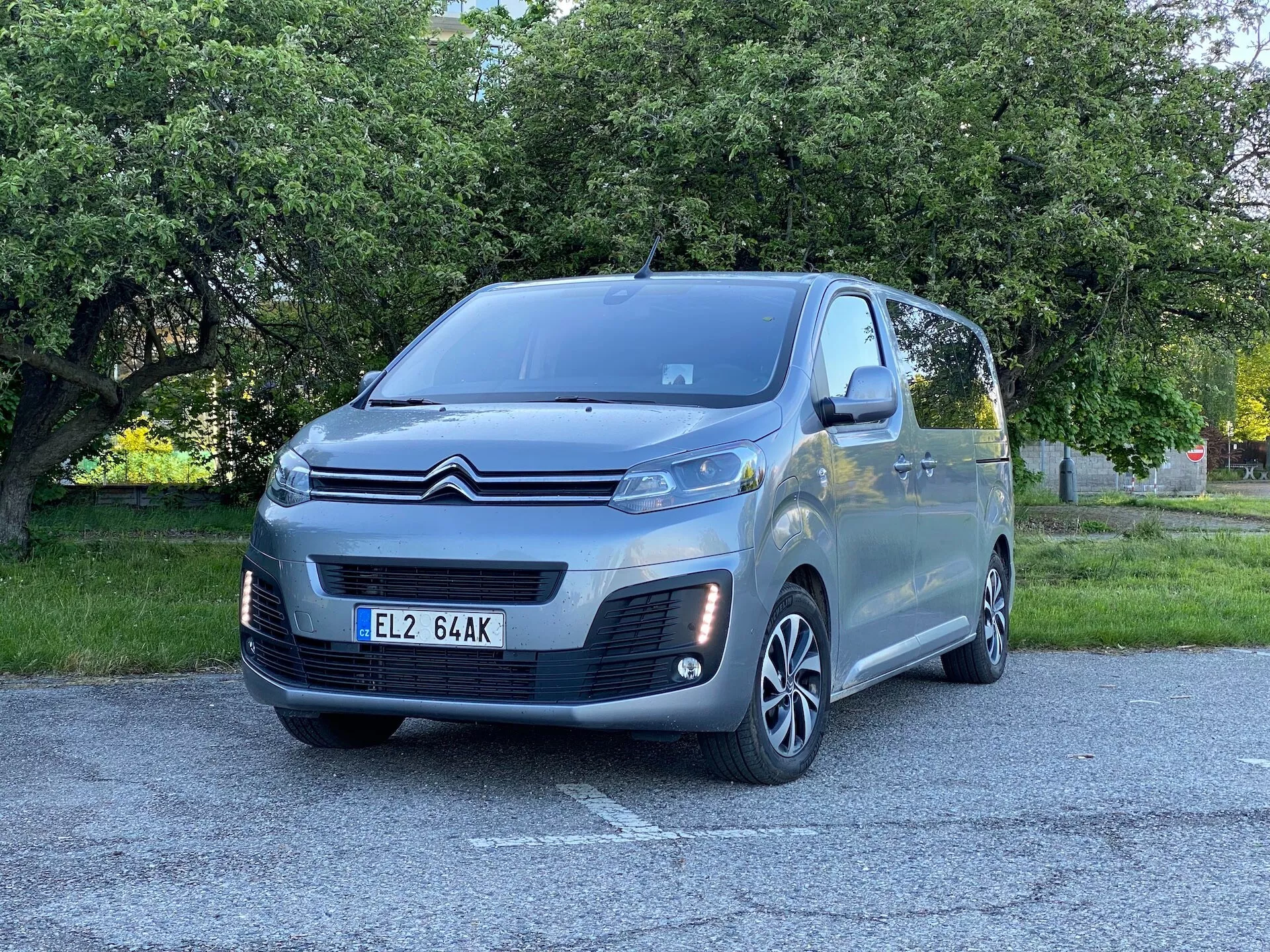 Obraz pokazuje szary minivan Citroën Spacetourer 2016 z trzema czwartymi widoku skierowanego na przednią lewą stronę pojazdu. Samochód jest zaparkowany na zewnątrz na utwardzonej powierzchni z zielenią, prawdopodobnie w miejskim parku lub na ulicy obsadzonej drzewami. Widoczne są kluczowe cechy: emblemat Citroëna w kształcie chevronu na czarnej i chromowanej przedniej kratce, nowoczesny design reflektorów, chromowane akcenty na światłach przeciwmgielnych, wieloszprychowe felgi aluminiowe, lusterka boczne w kolorze nadwozia z wbudowanymi kierunkowskazami, duże okna z przyciemnieniami na tylnych rzędach, wysoki dach typowy dla minivanów i prowadnica drzwi przesuwnych dla pasażerów z tyłu. Fotografia uchwyca te szczegóły z niskiego do średniego kąta, blisko samochodu, podkreślając teksturę nadwozia i styl kół.