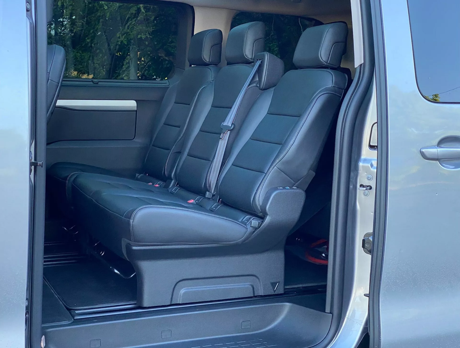 Wnętrze Citroëna SpaceTourer 2016 ze szczególnym uwzględnieniem miejsc pasażerskich. Zdjęcie wykonane pod kątem ukośnym przy tylnych drzwiach pasażera, ukazujące drugą i trzecią rząd siedzeń. Drugi rząd składa się z dwóch indywidualnych czarnych skórzanych foteli, podczas gdy trzeci rząd posiada ławkę dla trzech pasażerów. Cechy obejmują zagłówki, czerwone klamry pasów bezpieczeństwa, złożony środkowy podłokietnik, kratkę głośnika na drzwiach, wentylację klimatyzacji, uchwyty na kubki i prowadnice w podłodze do regulowanych siedzeń.