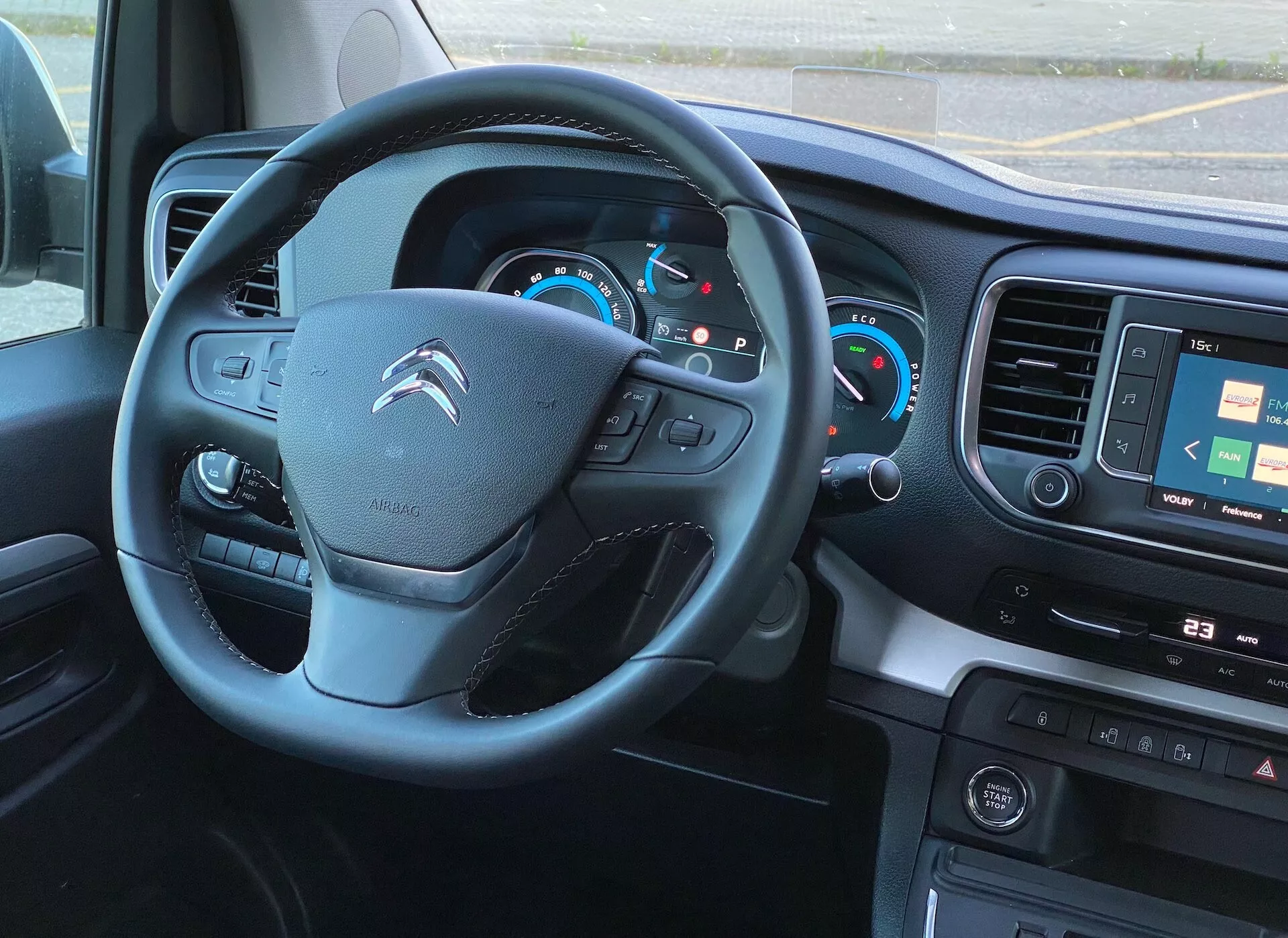 Widok wnętrza z perspektywy fotela kierowcy nowoczesnego Citroëna Spacetourer 2016, z naciskiem na kierownicę i deskę rozdzielczą. Kierownica z zamontowanymi kontrolkami sugeruje integrację systemów audio, łączności telefonicznej i regulacji tempomatu. Podświetlony panel instrumentów zawiera analogowe prędkościomierze i obrotomierze z centralnym wyświetlaczem cyfrowym. Ekran multimedialny pokazujący mapę, fizyczne przyciski do sterowania mediami i ustawieniami pojazdu oraz sterowanie klimatyzacją wskazują na zaawansowane funkcje technologiczne i komfortowe. Widoczny jest również panel sterowania z przyciskami wspomagania kierowcy. Zdjęcie jest zrobione z lekko podwyższonego kąta, ukazując dobrze oświetlone wnętrze z naturalnym światłem.