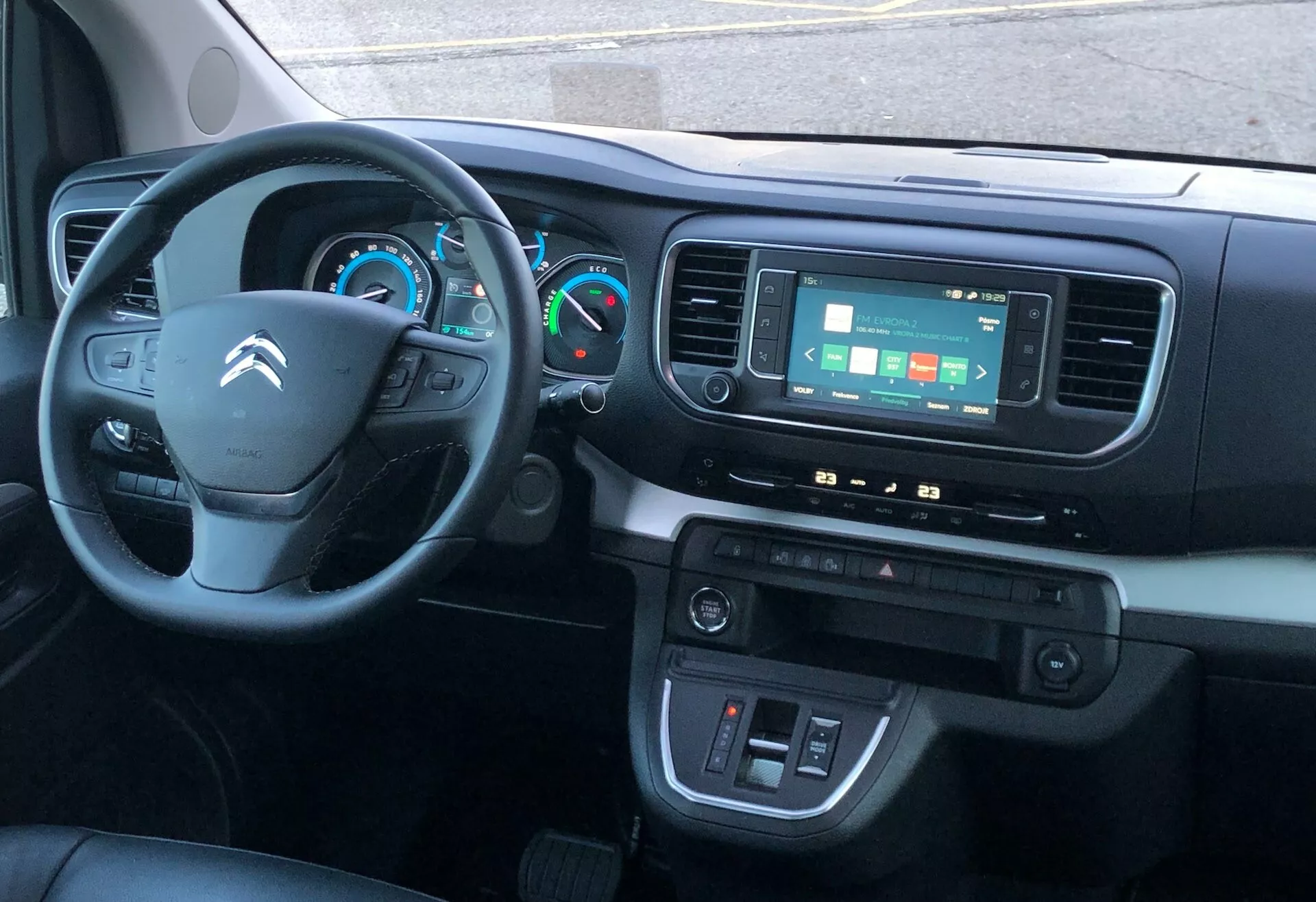 Widok wnętrza Citroëna SpaceTourer 2016 z przedniego siedzenia pasażera, ukazujący deskę rozdzielczą, trójramienną kierownicę z przyciskami, częściowo widoczny panel instrumentów z analogowymi i cyfrowymi wskaźnikami, system infotainment z ekranem dotykowym, pokrętła klimatyzacji oraz drzwi kierowcy z kontrolkami okien i lusterek bocznych. Środkowa konsola najprawdopodobniej zawiera przestrzeń do przechowywania i porty pomocnicze.
