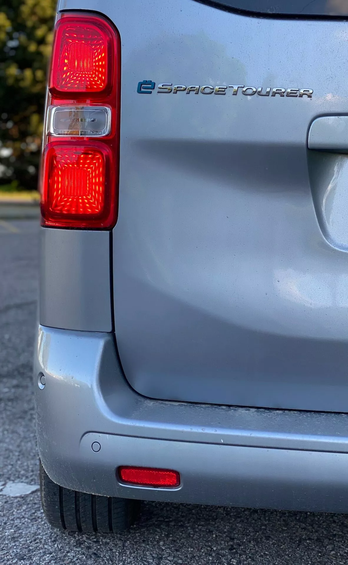 Zbliżenie tylnej części elektrycznego samochodu Citroen e-SpaceTourer z 2016 roku, wykonane z niskiego do średniego kąta, ukazujące lewy zespół świateł tylnych z pionowymi czerwonymi światłami i zintegrowanymi czujnikami parkowania w zderzaku, na tle słonecznego nieba, podkreślające srebrną farbę i lekkie zużycie powierzchni pojazdu.