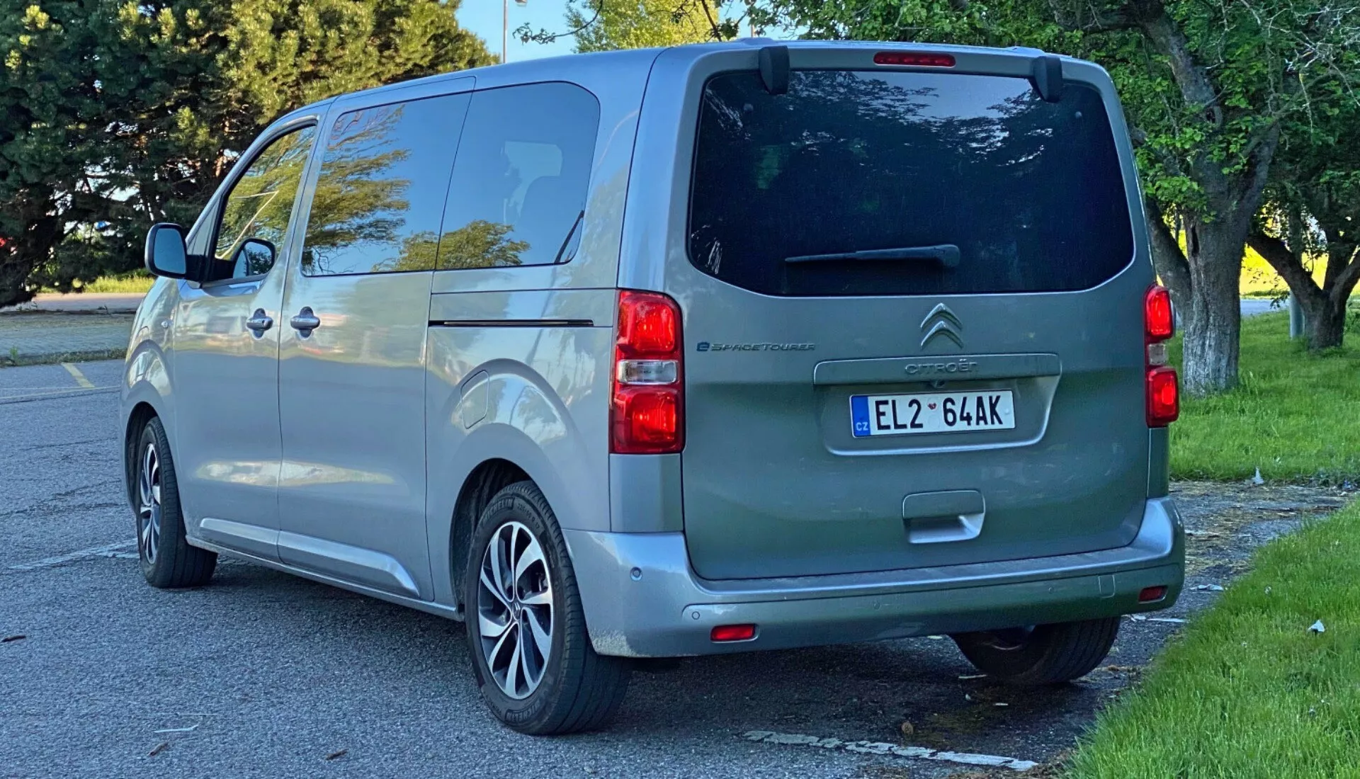 Tylny widok metalicznie szarego vana Citroën SpaceTourer 2016, uchwycony pod kątem trzech czwartych, podkreślający lewą stronę, ukazujący klapę bagażnika, tylną szybę, dwukolorowy zderzak z czujnikami parkowania i podświetlony lewy klaster świateł. Przesuwane drzwi vana z widoczną szyną nad tylnym kołem i alufelgi sugerują kombinację stylu i funkcjonalności, w miejskim otoczeniu sugerującym światło dzienne, prawdopodobnie po południu. Przyciemnione okno dla prywatności i ogólna standardowa konfiguracja podkreślają przydatność pojazdu do użytku osobistego, rodzinnego lub małych firm.
