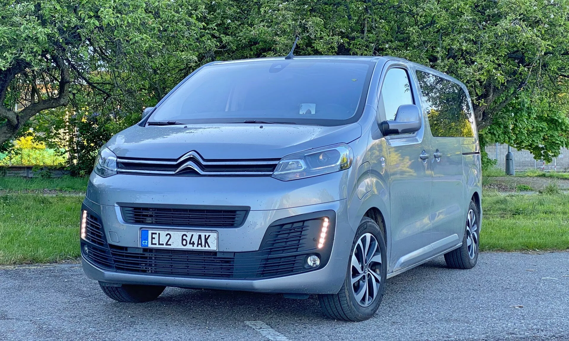 Obraz przedstawia nowoczesny Citroen SpaceTourer 2016 ustawiony pod kątem, z przodem zwróconym lekko w prawo. Widoczna jest strona kierowcy, pełny przód i fragment bocznego profilu. Samochód zajmuje dużą część kadru, podkreślając swoje reflektory LED, charakterystyczną przednią kratkę, wieloramienne felgi aluminiowe, lusterka boczne z kierunkowskazami i przednie czujniki parkowania. Metaliczne wykończenie i elegancki design podkreślają nowoczesną inżynierię motoryzacyjną. Scena rozgrywa się na świeżym powietrzu w parku w ciągu dnia, charakteryzując się naturalnym oświetleniem i drzewami oraz trawą w tle.