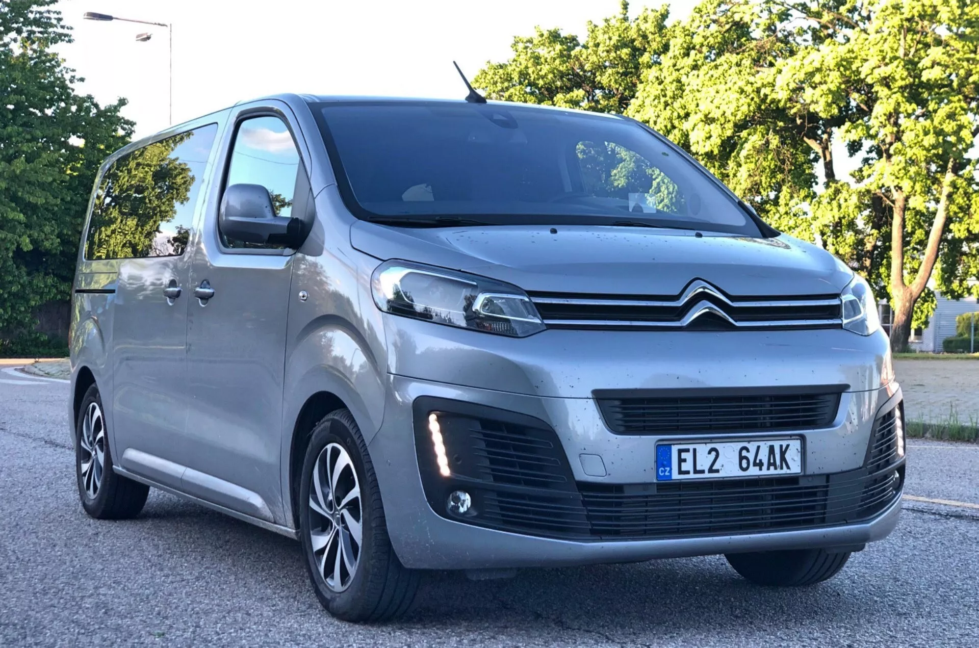 Nisko kątowy, trzy-czwarte widok z przodu metalicznego szarego Citroëna Spacetourer MPV 2016, podkreślający wypukły przedni grill z logo podwójnego chevronu, rzeźbione reflektory LED lub HID, czujniki parkowania na zderzaku, wieloramienne felgi aluminiowe, aerodynamiczne lusterka boczne z wbudowanymi kierunkowskazami i duże okna z rosnącą linią okien. Samochód jest zaparkowany na asfaltowej powierzchni w podmiejskim lub parkowym otoczeniu z liśćmi w tle.