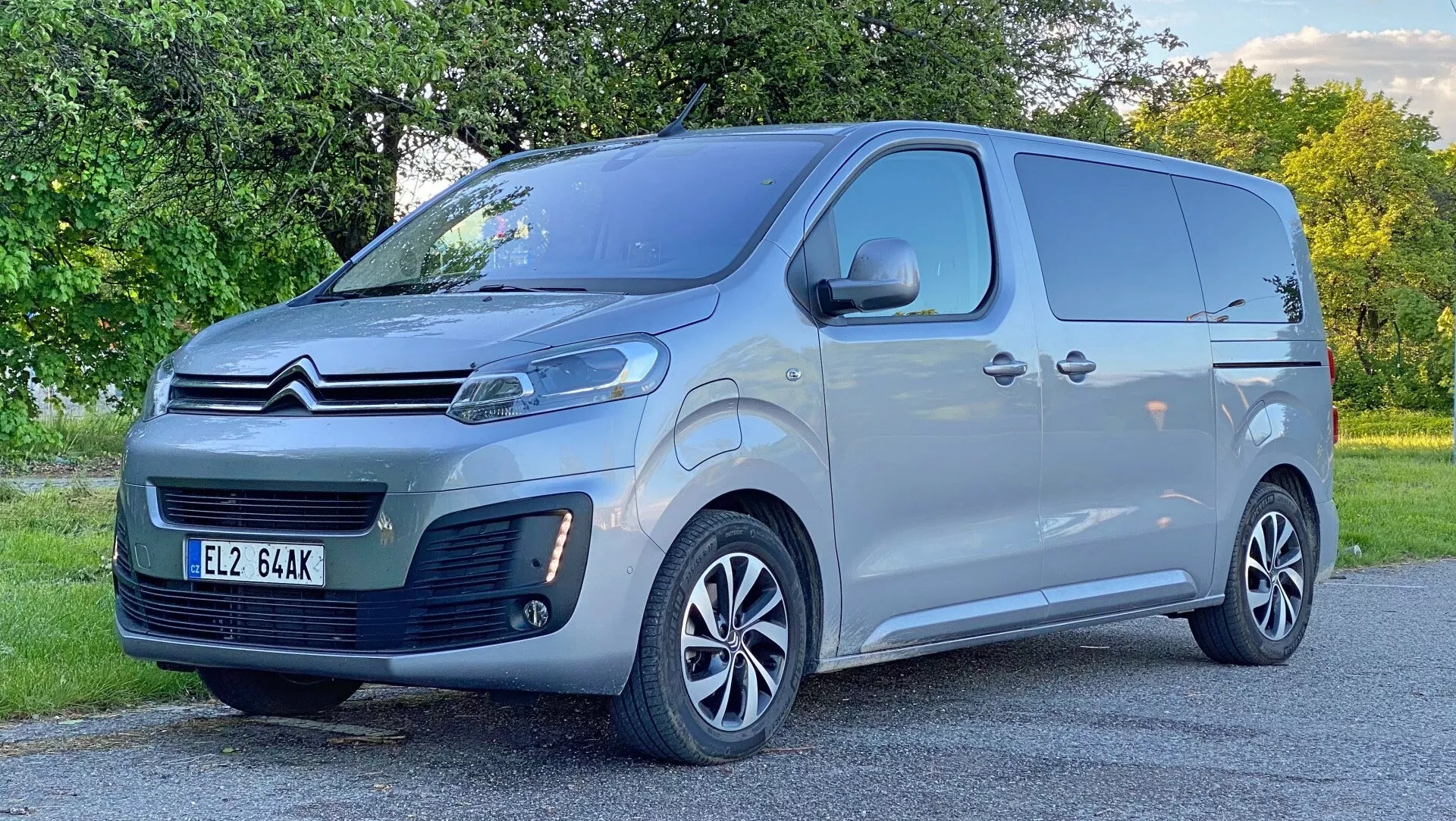 Zdjęcie furgonu Citroën Spacetourer 2016 wykonane z niskiego przedniego trzykwartego kąta, pokazujące jego przedni i boczny profil. Nowoczesny, elegancki design zawiera wyraźne podwójne logo Citroëna na kratce, charakterystyczne reflektory prawdopodobnie z dziennymi światłami LED, wyśrodkowaną przednią tablicę rejestracyjną i dolny wlot powietrza na przednim zderzaku. Pojazd jest pomalowany na jasnoszary kolor, ma duże lusterka boczne i wieloramienne felgi aluminiowe, co wskazuje na średni lub wyższy poziom wyposażenia. Boczne profile obejmują przesuwne drzwi po stronie kierowcy i solidne sekcje paneli do przewozu towarów. Tło to przydrożna zieleń, uchwycona przy naturalnym świetle.