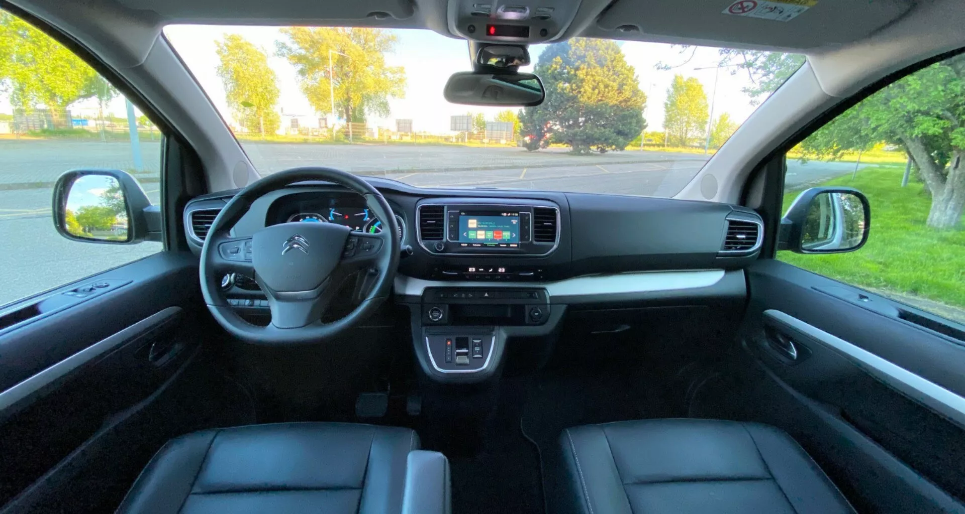 Widok wnętrza Citroen Spacetourer 2016 z perspektywy tylnego siedzenia, ukazujący kierownicę z zintegrowanymi elementami sterującymi, cyfrowy panel instrumentów, konsolę środkową z wyświetlaczem infotainment, przyciski sterowania klimatyzacją, dźwignię zmiany biegów, elektroniczny przycisk hamulca postojowego i elementy sterujące drzwiami. Nowoczesna technologia motoryzacyjna i ergonomia są widoczne z ozdobnymi elementami z szczotkowanego metalu lub drewna oraz skoordynowaną kolorystyką.