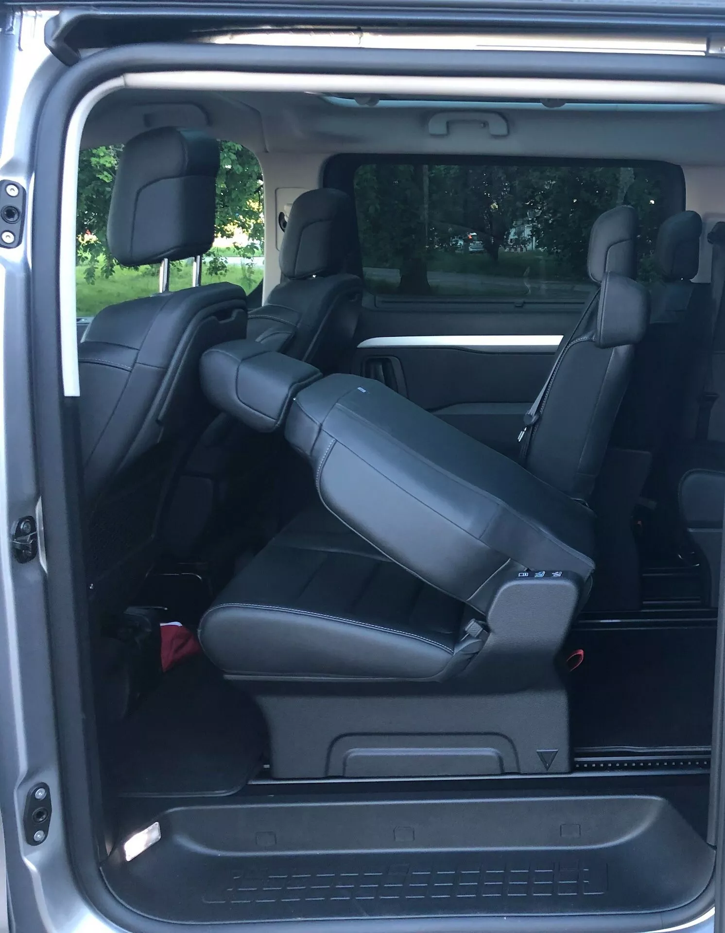 Widok wnętrza Citroena Spacetourer 2016 z perspektywy tylnej, koncentrujący się na tylnych siedzeniach. Nowoczesny minivan posiada trzy rzędy siedzeń. Środkowy rząd, z fotelami kapitańskimi, jest nachylony do przodu, pokazując funkcję składania w celu zwiększenia przestrzeni ładunkowej lub dostępu do trzeciego rzędu. Tylna rząd obejmuje dwa siedzenia z zagłówkami i dużą przestrzeń na nogi. Wnętrze wyłożone jest czarną skórą dla luksusowego wrażenia, pasy bezpieczeństwa oraz otwór wentylacyjny na pionowym słupku, co wskazuje na funkcje klimatyzacji tylnej.